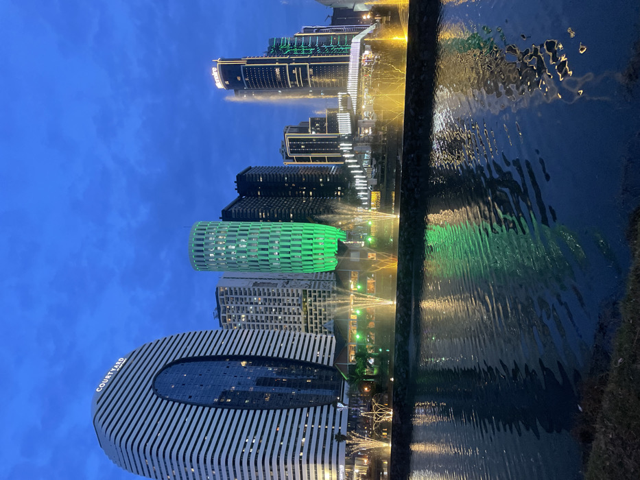 The image depicts a modern cityscape at dusk or night, viewed across a body of water. The scene is illuminated by artificial lights from numerous buildings and fountains, creating vivid reflections on the rippling water surface.
In the mid-ground, a dark body of water, possibly a canal or bay, spans the width of the frame. A dividing wall separates the water from the foreground, where several luminous fountains project jets of water into the air. These fountains are lit in shades of yellow and green, casting vibrant glows onto the water and the surrounding area. The water itself reflects the colorful lights of the distant buildings, creating a dynamic interplay of light and shadow.
The background is dominated by a cluster of contemporary skyscrapers. On the far left, a prominent, curved white building features vertical linear patterns and a large oval dark glass facade. The text "COURTYARD" is visible in white letters on its upper left side. Adjacent to it are other tall structures, including a distinctive cylindrical building with a bright green illuminated facade. Further to the right, a series of dark skyscrapers with illuminated windows and linear architectural lighting contribute to the urban skyline. In the far distance, more buildings with intricate light patterns can be seen.
The sky above is a deep blue, characteristic of twilight or early night, with hints of clouds. No specific human activity is depicted; the scene captures the static beauty of an illuminated urban landscape. Based on the architectural style and the visible text "COURTYARD," the location is Batumi, Georgia, featuring its modern architectural developments.