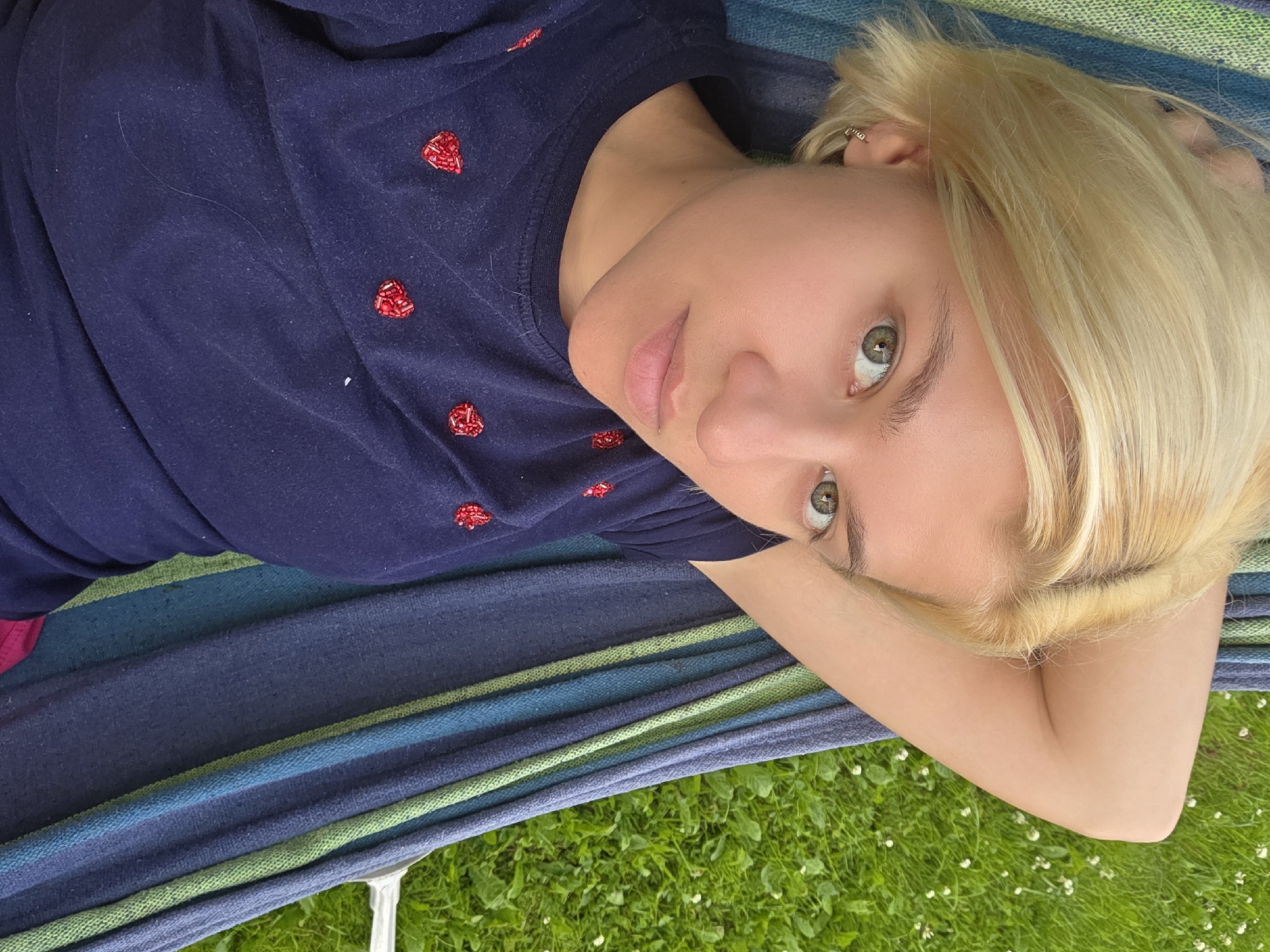 A young woman with blonde hair and green eyes is lying on her back in a hammock. She is wearing a dark blue t-shirt adorned with small, beaded red hearts. Her head is propped up on her arm, and she is looking directly at the camera. Her expression is relaxed and pleasant. The hammock itself is made of striped fabric in shades of blue, green, and purple. Below the hammock, a patch of green grass with small white flowers is visible, suggesting an outdoor setting. The lighting suggests it is daytime. The image was taken in Frolovo, Russia.