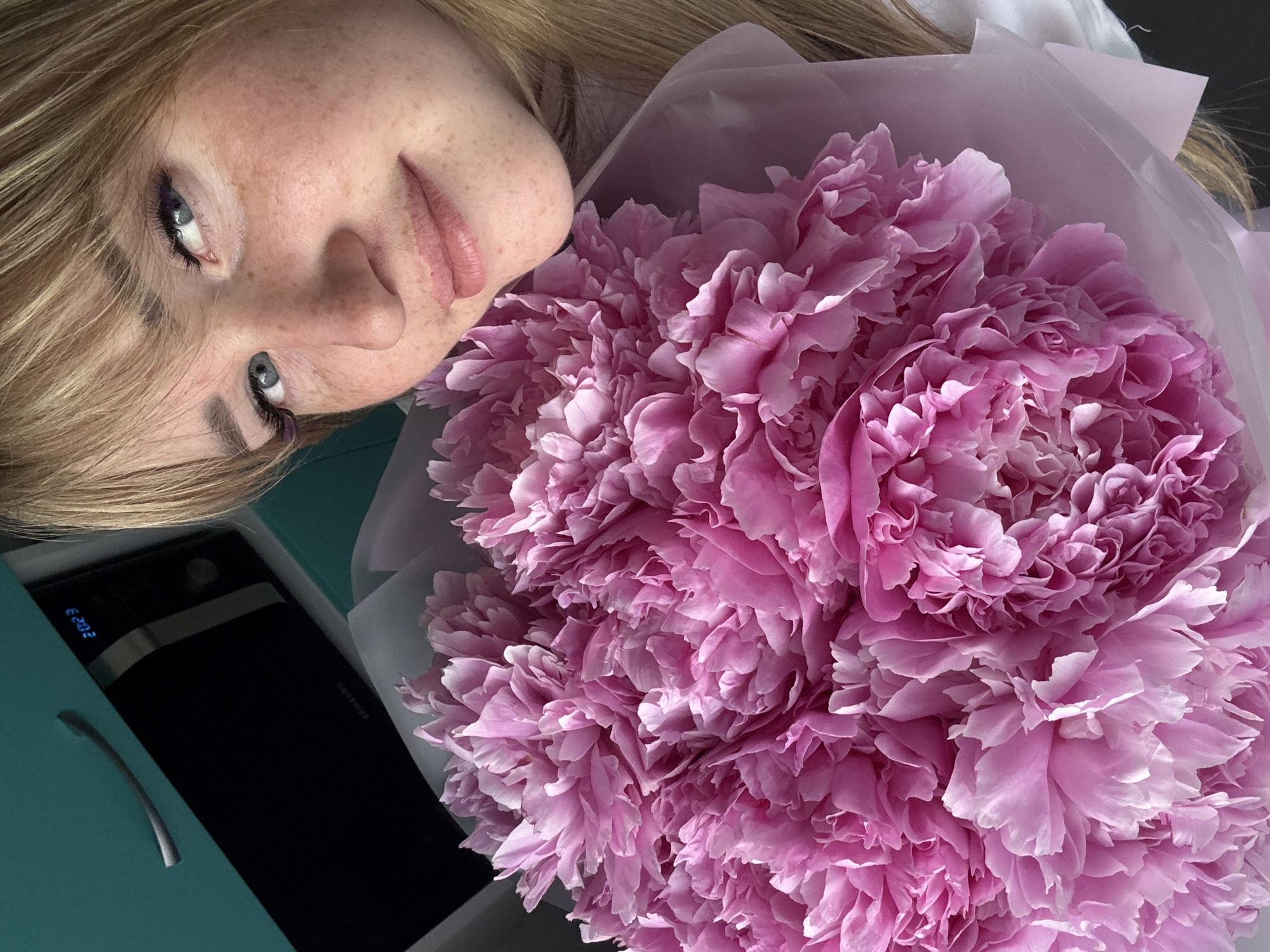 A young woman with blonde hair and freckles is lying on her side, looking at the camera. She has blue eyes and is wearing makeup, including eyeliner. Beside her is a large bouquet of pink peonies, wrapped in light pink paper. In the background, a black electronic device with a digital display showing "E 202" is visible. The scene appears to be indoors, possibly in a bedroom or living area. The lighting suggests it might be daytime. The overall mood is peaceful and pleasant.