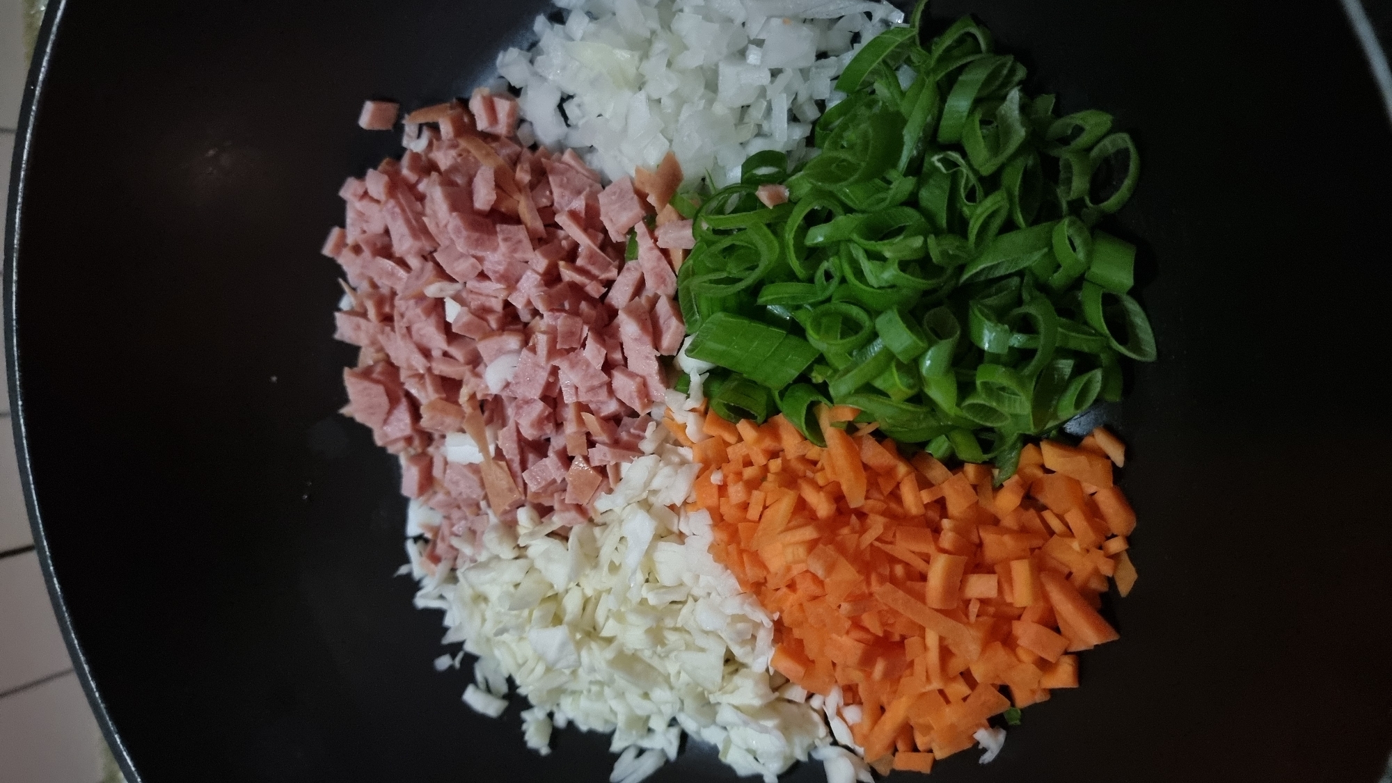 The image shows a black wok containing chopped ingredients organized into four sections. The top left quadrant features diced ham, transitioning into chopped white onion in the top right. The bottom right quadrant contains diced carrots, while the bottom left quadrant holds sliced green onions. The wok sits on a surface with what appears to be a light-colored tile pattern visible along the left edge. The overall arrangement suggests a preparation for a stir-fry or similar dish. There is no information indicating the location is Dunnae-myeon, South Korea.