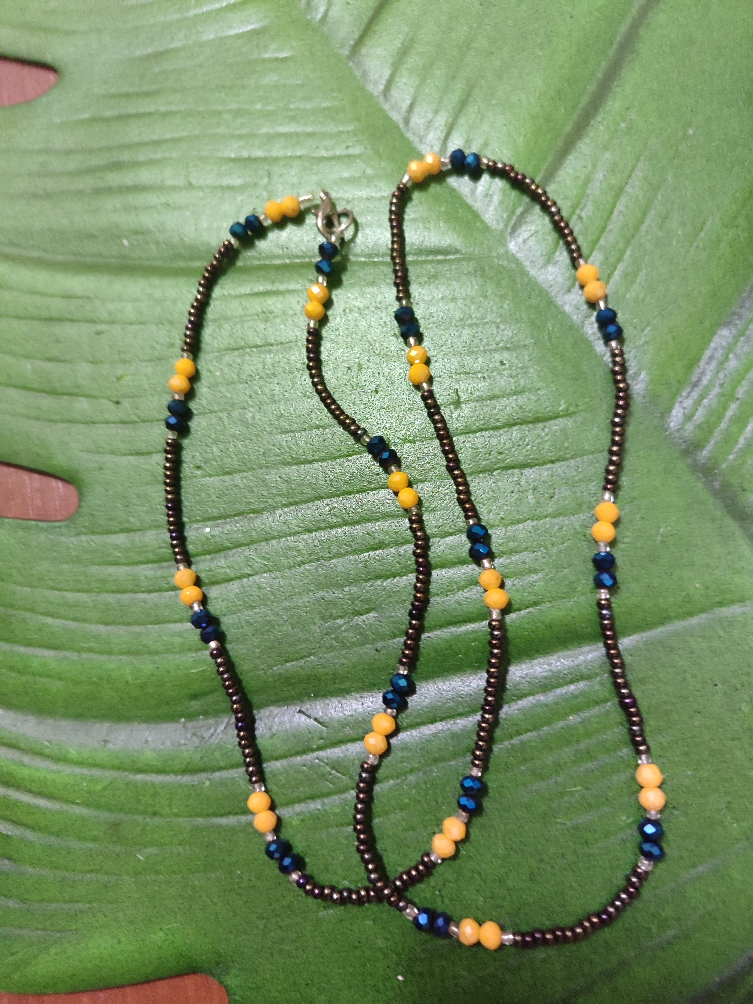 The image shows a close-up, overhead view of a beaded necklace lying on a large green leaf. The necklace is composed of small, dark brown beads with occasional clusters of yellow and blue faceted beads. The necklace has a clasp closure visible at the top left of the frame. The leaf has prominent veins and a slightly textured surface. The background appears to be a wooden surface, visible in small areas at the bottom and left edges of the image. No people or specific locations within the city of Mykolaiv, Ukraine are identifiable in this image.