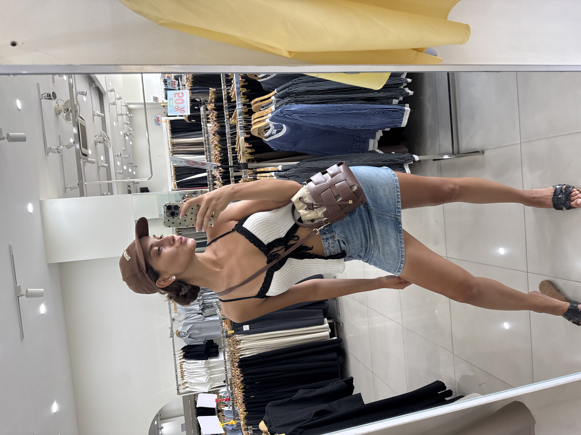 A woman is seen in a mirror selfie in a clothing store. She is wearing a brown cap, a white top with black trim, a denim skirt, and sandals. She is holding a phone in her left hand and a brown woven purse is slung over her shoulder. The background shows clothing racks with various garments in black, white, and blue. The store appears to be well-lit. The image does not provide information about the city of Đà Nẵng, Vietnam.