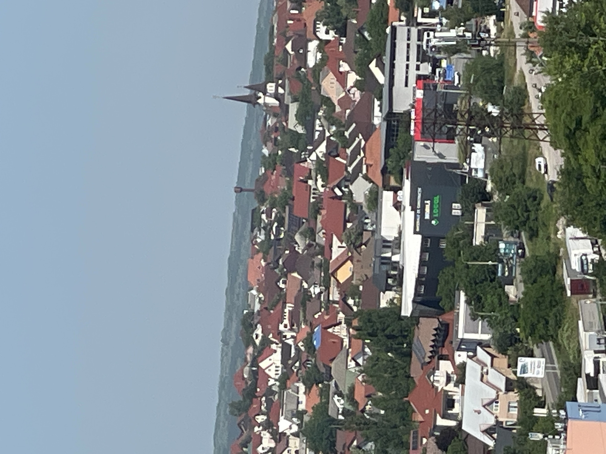 The image displays a sprawling residential area with numerous houses featuring red-tiled roofs, set against a backdrop of rolling hills under a clear blue sky. In the mid-ground, a prominent church with twin spires stands out, while a water tower is visible in the distance. Below the residential section, several commercial buildings are present, including one with the visible text "LOGOL". Various vehicles, including trucks and cars, are scattered throughout the scene, and electrical pylons are also noticeable. The overall impression is of a moderately sized town or city.