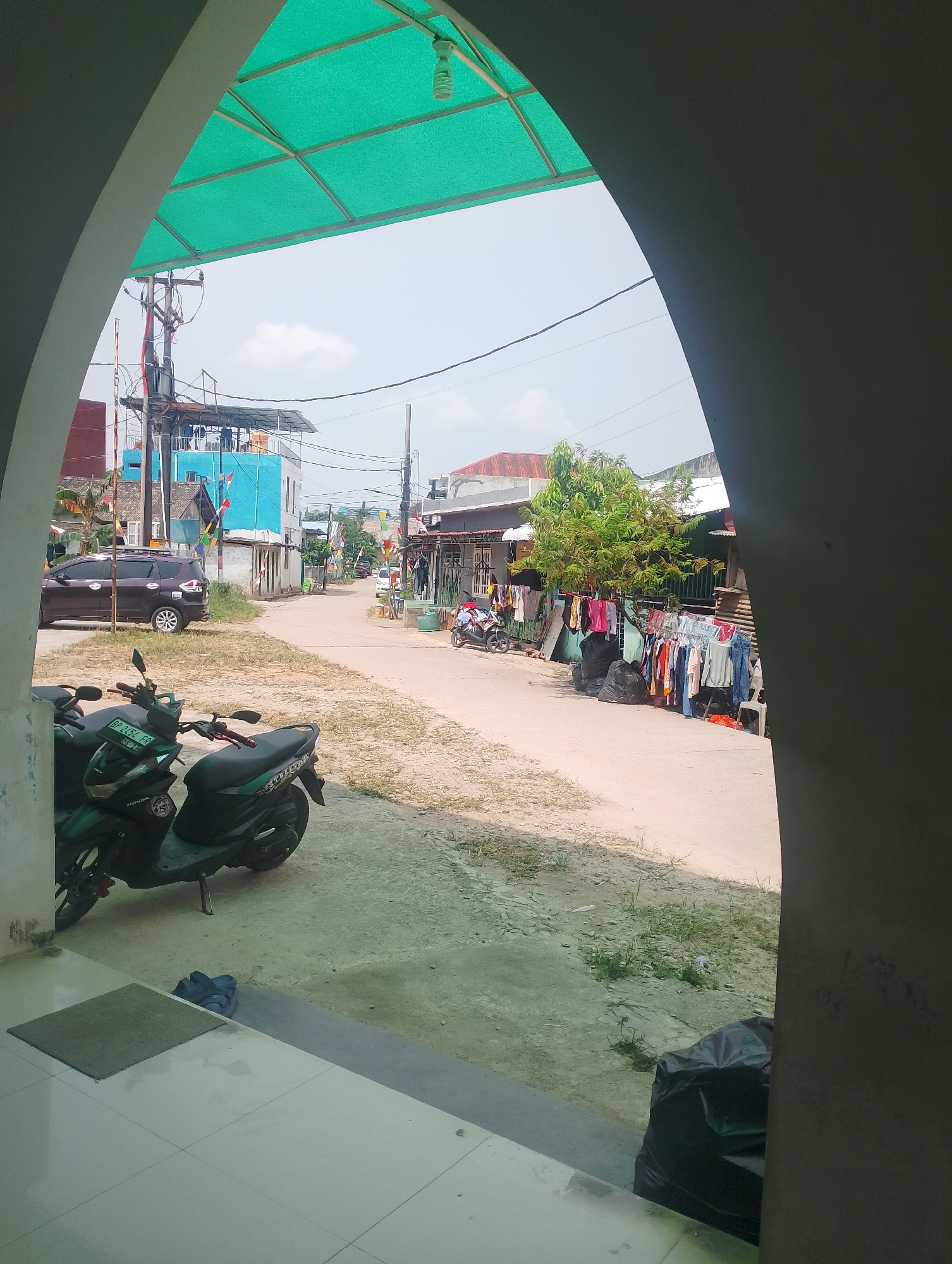The image captures a street scene in Batam, Indonesia, as viewed through an archway. The foreground features a tiled floor with a doormat and a pair of blue sandals. Two green scooters are parked on the left side of the image, one with a license plate reading "BP 2452 EP". The street itself is paved and appears to be a residential or commercial area, with buildings on either side.
On the left side of the street, a dark-colored SUV is parked. Further down the street, a blue building stands prominently, with a transformer and utility poles nearby. Small flags, possibly decorative or national, are strung across the street.
On the right side of the street, laundry is hanging on a clothesline. Several black trash bags are also visible along the roadside. Trees and foliage are present, suggesting a tropical environment. The sky is bright, indicating daylight. The overall impression is one of a typical day in a bustling Indonesian neighborhood.