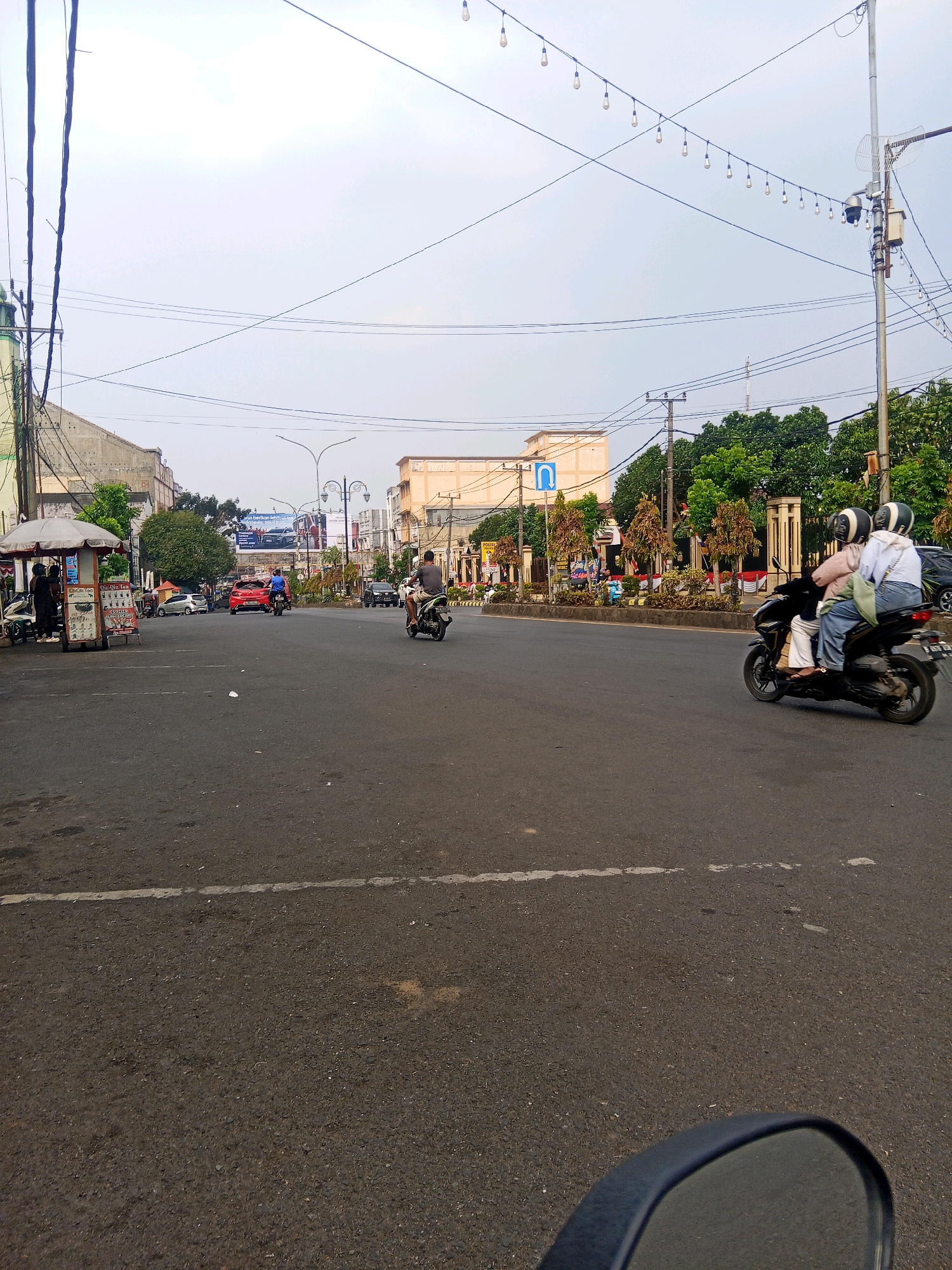 This image depicts a street scene in Lubuklinggau, Indonesia, likely near a busy intersection or commercial area. In the foreground, the asphalt road is visible with a white line marking. The middle ground shows a road with several vehicles, including two motorcycles. One motorcycle, in the right half of the image, carries two individuals wearing helmets, the passenger in the rear wearing a white long-sleeved shirt and light-colored pants, and the driver wearing a light pink top and dark pants. Another motorcycle is further down the road to the left. A red car is also present. On the left side of the road, there is a small roadside stall with an umbrella canopy and several people. A billboard displaying advertisements is visible in the background, along with buildings and trees. Overhead, numerous electrical wires crisscross the sky, strung between poles, with some decorative lights also suspended. The sky is overcast, indicating a cloudy day.