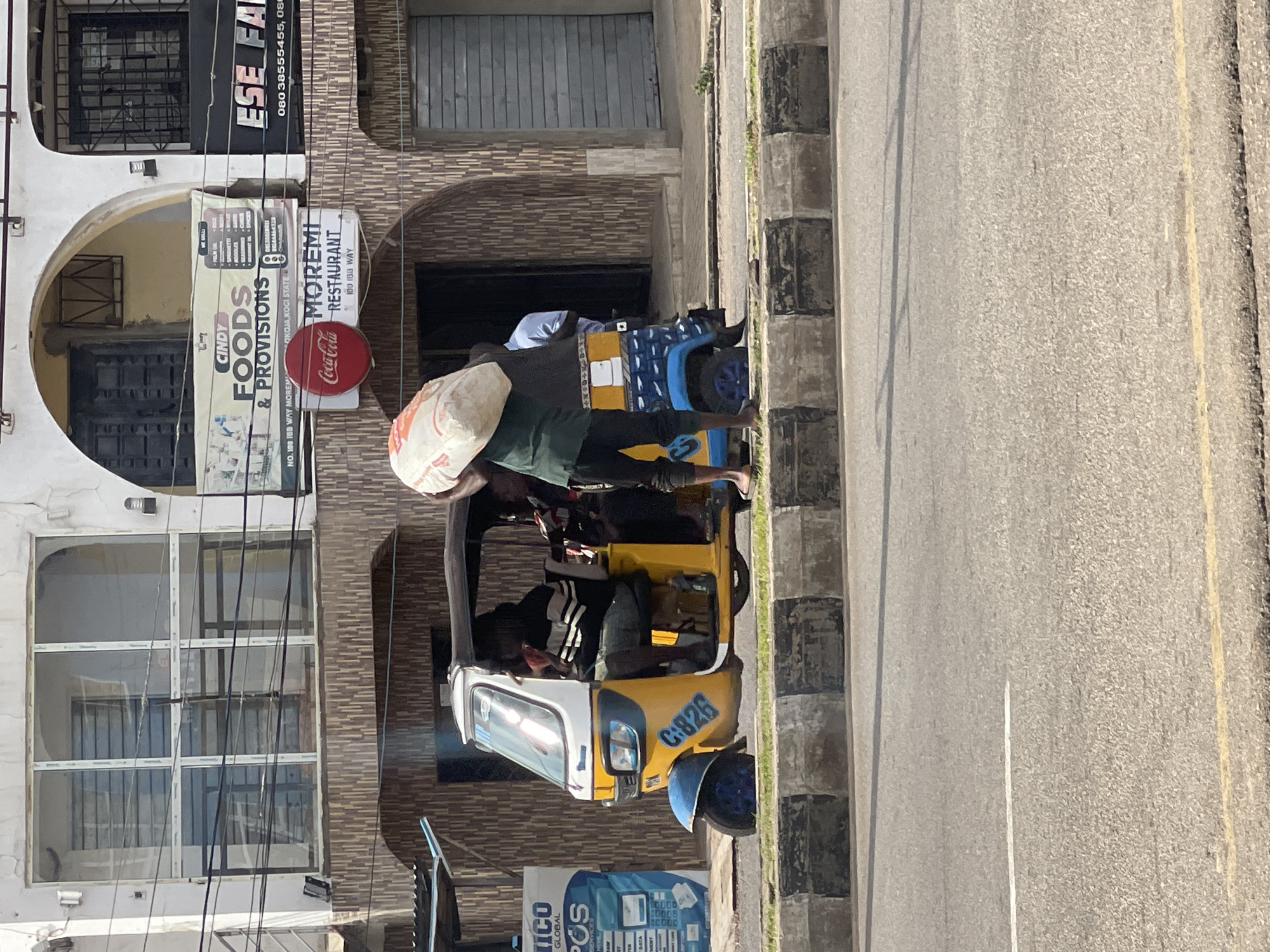 In the city of Unknown, Unknown, a person carrying a large sack of goods on their shoulders walks alongside a yellow and blue auto-rickshaw parked at the side of the road. The auto-rickshaw has a black canopy and two individuals are seated inside. A man in a striped shirt is in the driver's seat. The scene is set against a backdrop of a building with various shops, including "CINDY FOODS & PROVISIONS" and "MOREMI RESTAURANT". The building features tiled lower sections and arched windows. A concrete barrier with black and white markings separates the sidewalk from the asphalt road. The overall environment appears to be an urban street.