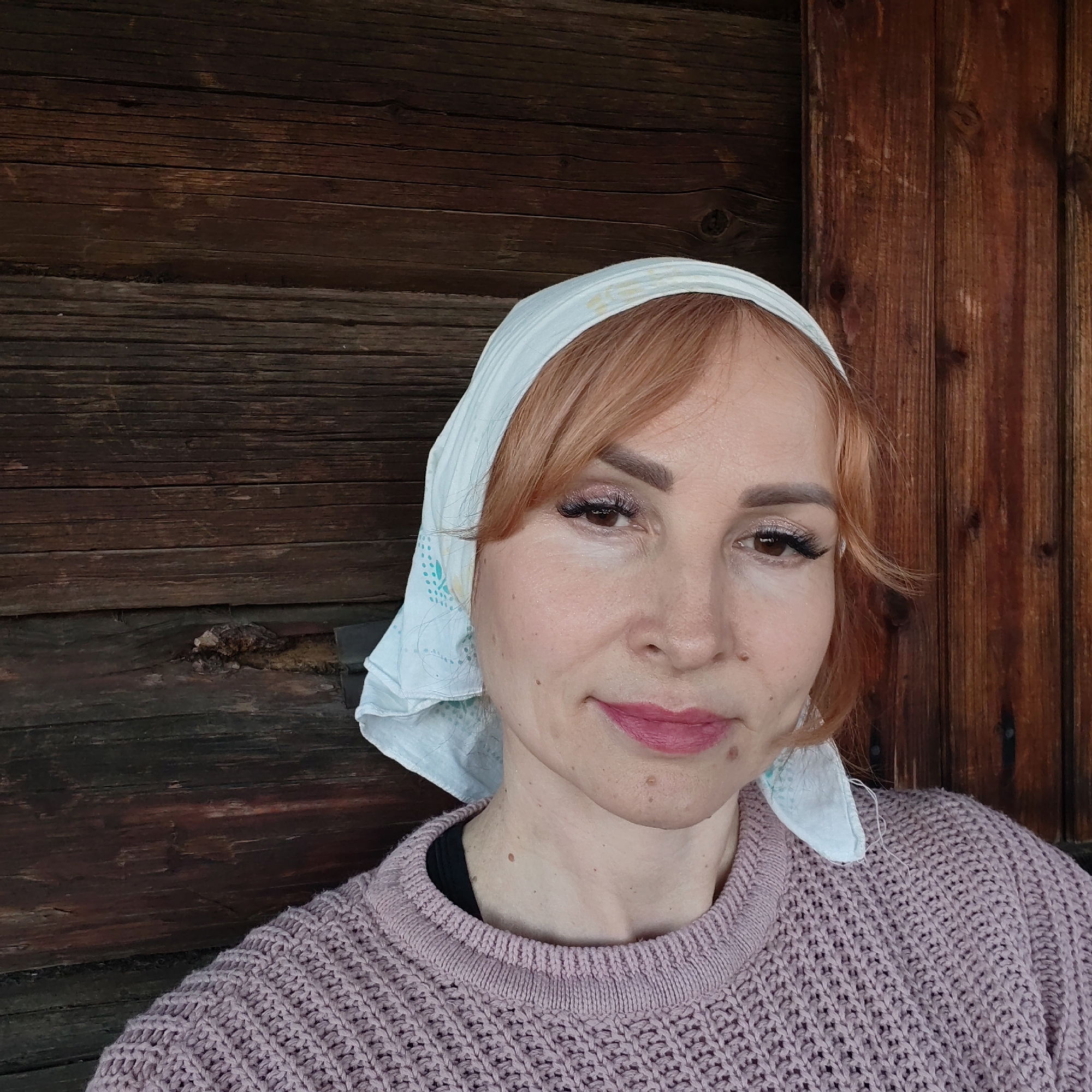 A middle-aged woman with red hair and a white headscarf poses for a selfie. She wears a pink knitted sweater and light makeup. The background is a rustic wooden wall with distinct horizontal planks. The location is indiscernible from the image alone, so a determination regarding Chaikovsky, Russia cannot be made.