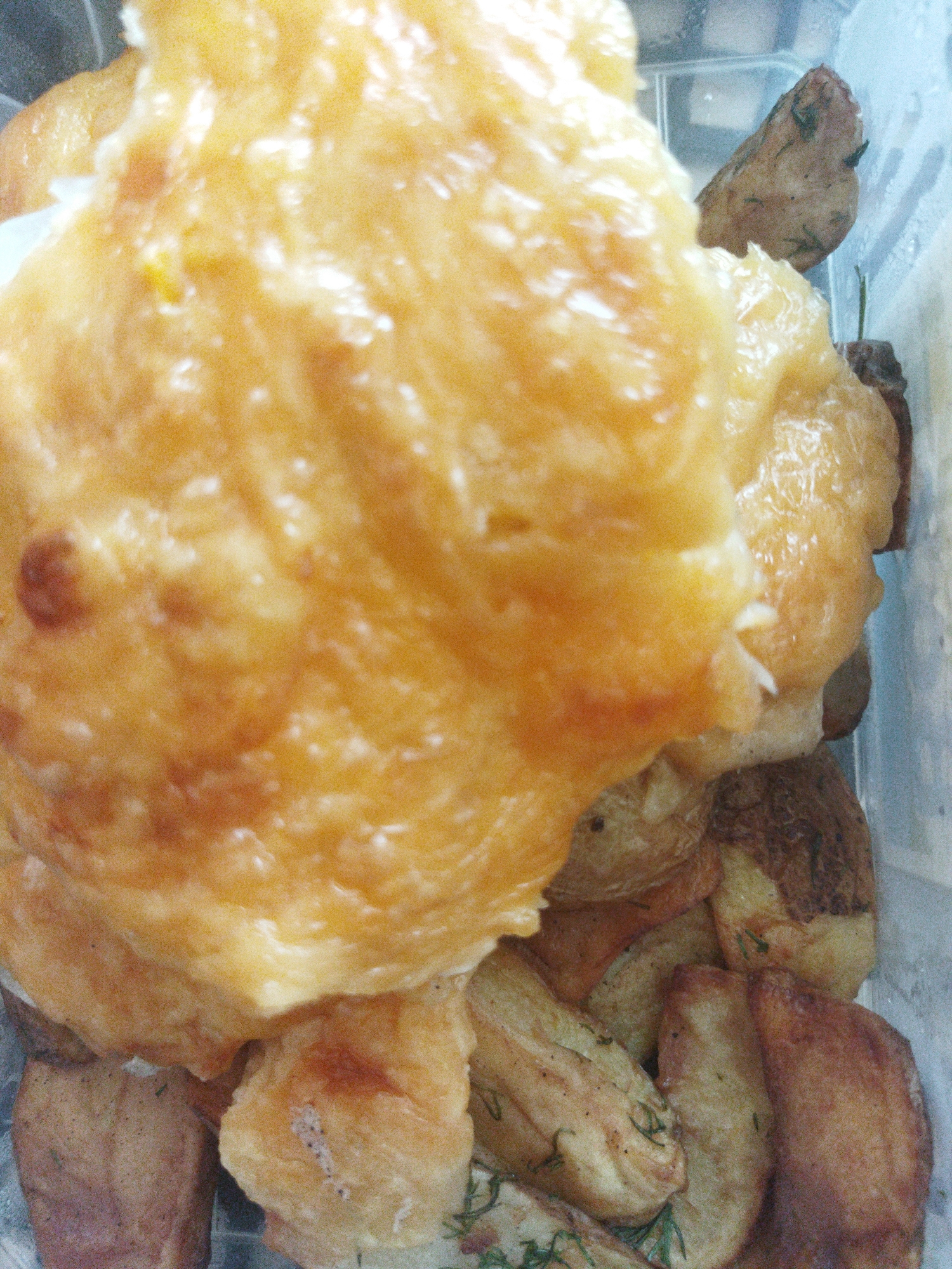 The image shows a close-up of food in a clear plastic container. The dominant feature is a large, golden-brown, shiny mass, possibly a type of cooked or baked doughy food, partially covering the contents below. Beneath this are potato wedges, some with herbs (likely dill), showing varying degrees of browning. The background is the inside of the container. There is no indication of Kramatorsk, Ukraine, or any other location-specific details. No people are visible.
