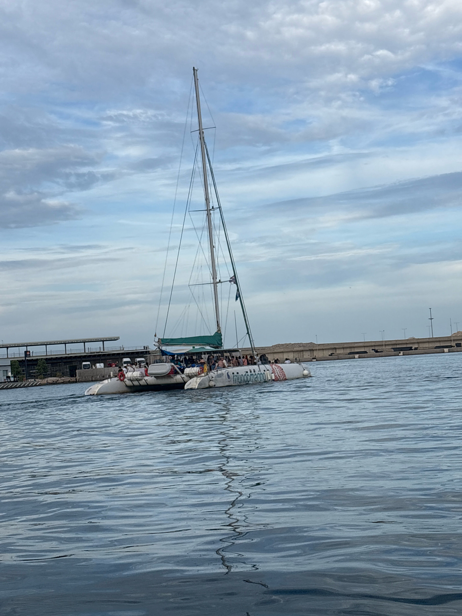 The image shows a catamaran sailing on a body of water, possibly the port of Valencia, Spain, given the presence of industrial structures and a bridge in the background. The catamaran is white with a green sail and has a group of people on board, appearing to be on a tour or leisure trip. The sky is overcast with some clouds. The water is calm with ripples, and a reflection of the catamaran is visible. On the port side of the catamaran, the text "mundomarine" is visible. A building complex with a covered walkway and some vehicles is visible on the left side of the image, suggesting a port or marina area. A concrete barrier and a bridge structure are visible in the distance on the right.