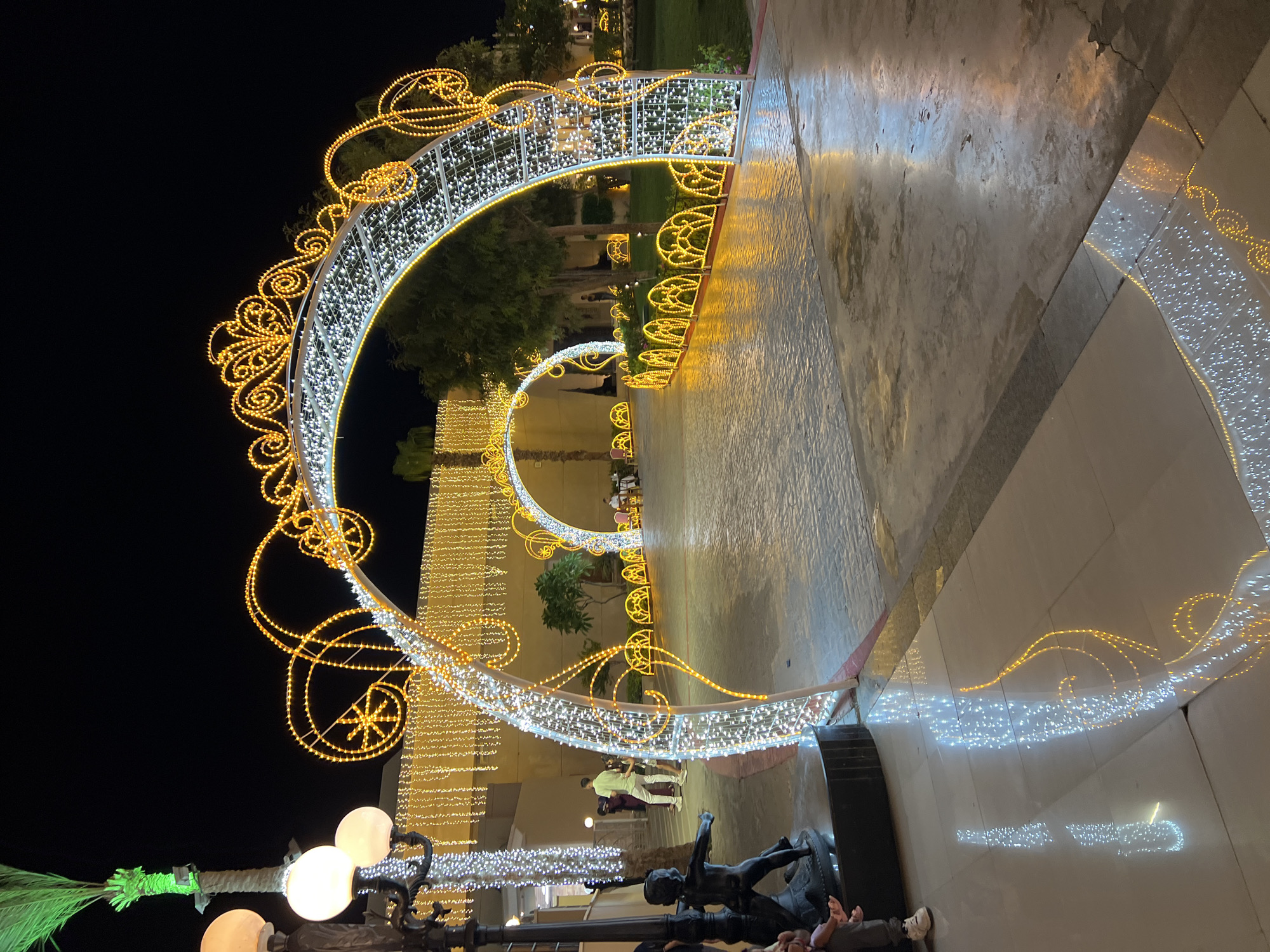This is a nighttime, ground-level, wide-angle photograph taken in Sharm El Sheikh, Egypt, at a decorative walkway. The scene is dominated by ornate archways adorned with white and gold string lights, creating a festive atmosphere. These archways lead into a dimly lit pathway lined with trees and illuminated by further string lights and decorative illuminated barriers.
In the foreground, to the left, a statue of a child is visible, partially obscured. A person is seated on the base of the statue, wearing gray pants and white sneakers. Further back on the left, two individuals are walking away from the camera; one is wearing a light green shirt and white pants, and the other is wearing a dark top and possibly a skirt.
The walkway itself is paved with textured tiles and is wet in places, reflecting the overhead lights. To the right, a well-maintained grassy area is visible, with more trees and illuminated decorative elements. The background is dark, indicating it is nighttime, with some buildings and further decorative lighting faintly visible. The overall impression is one of a beautifully illuminated outdoor space, likely a resort or entertainment area in Sharm El Sheikh.