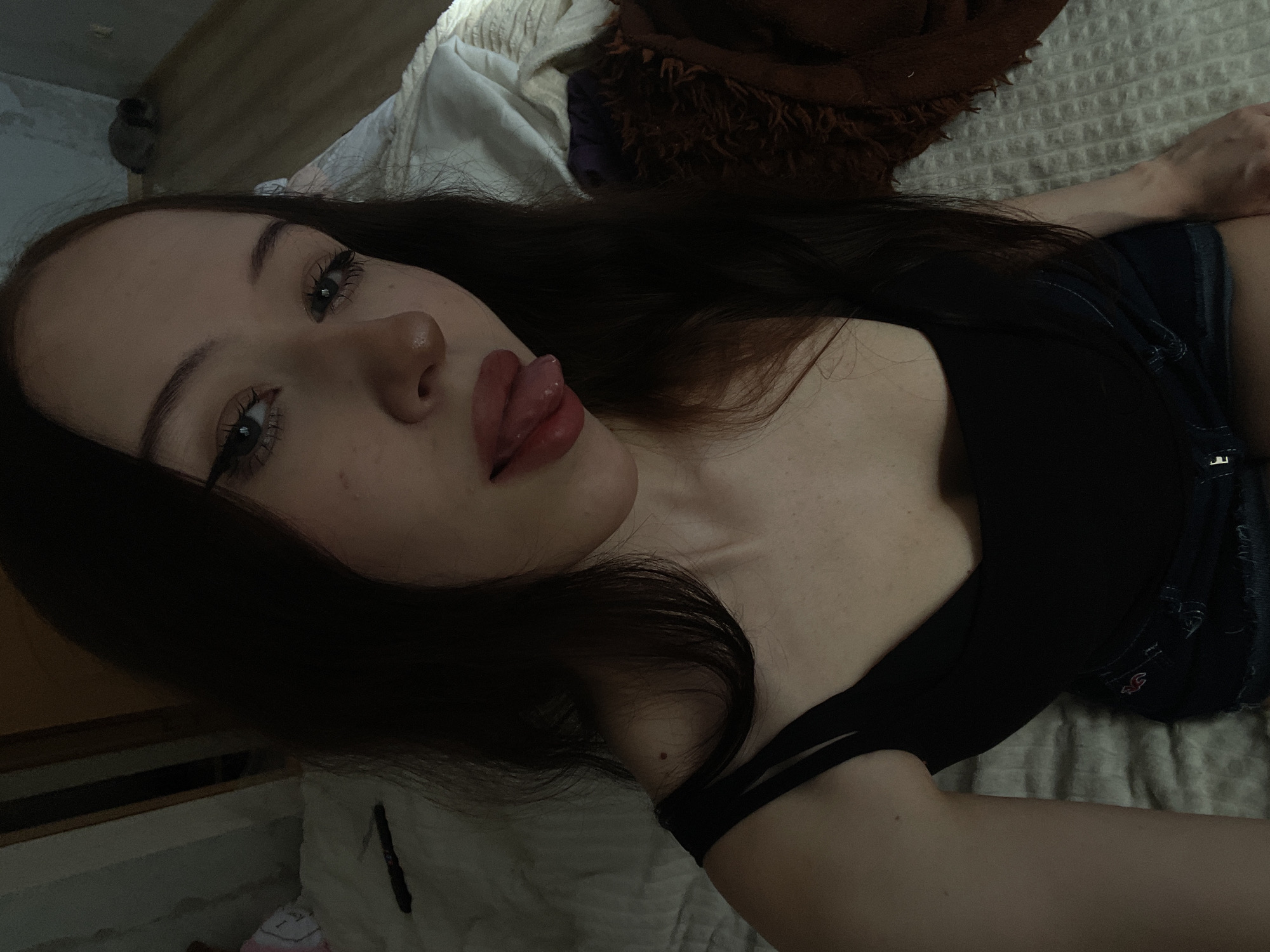 The image shows a close-up, indoor selfie of a young woman. She has long, dark hair and is wearing a black tank top and denim shorts. Her tongue is sticking out slightly. The background appears to be a bedroom, with a bed and some bedding visible. The lighting is dim. There is no discernible information in the image that would allow for identification of the location within the city of Votkinsk, Russia.