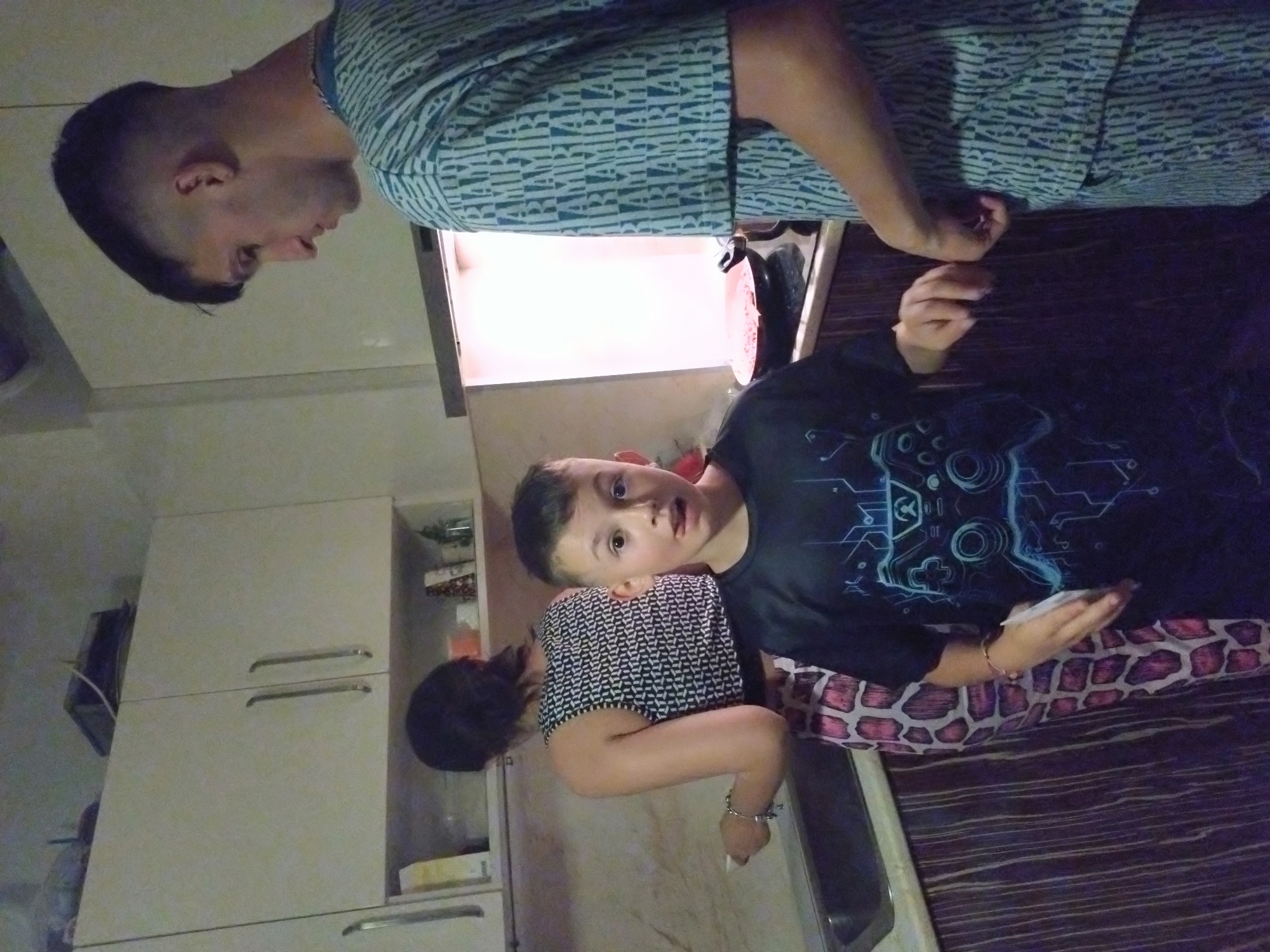 The image depicts three individuals in what appears to be a kitchen. A young boy wearing a dark t-shirt with a game controller design is lying down, holding a white object in his hand. A woman with dark hair, wearing a black and white patterned top, is standing beside him. A man in a light blue patterned shirt is standing above them. The kitchen includes white cabinets with handles and what seems to be a sink. The image provides no information that allows locating it in the city of Stara Zagora, Bulgaria.