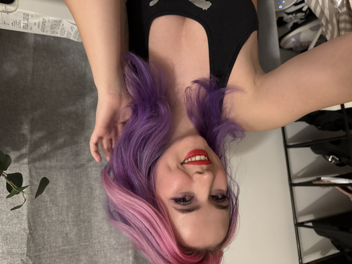 A woman with purple and pink hair, red lipstick, and eyeshadow smiles at the camera. She wears a black tank top. Her right hand is raised near her face. To the left is a gray textured surface with a green leafy stem. Behind her on the right, is a white wall and a metal shoe rack. Sneakers are visible in the background. The image is rotated counter-clockwise.