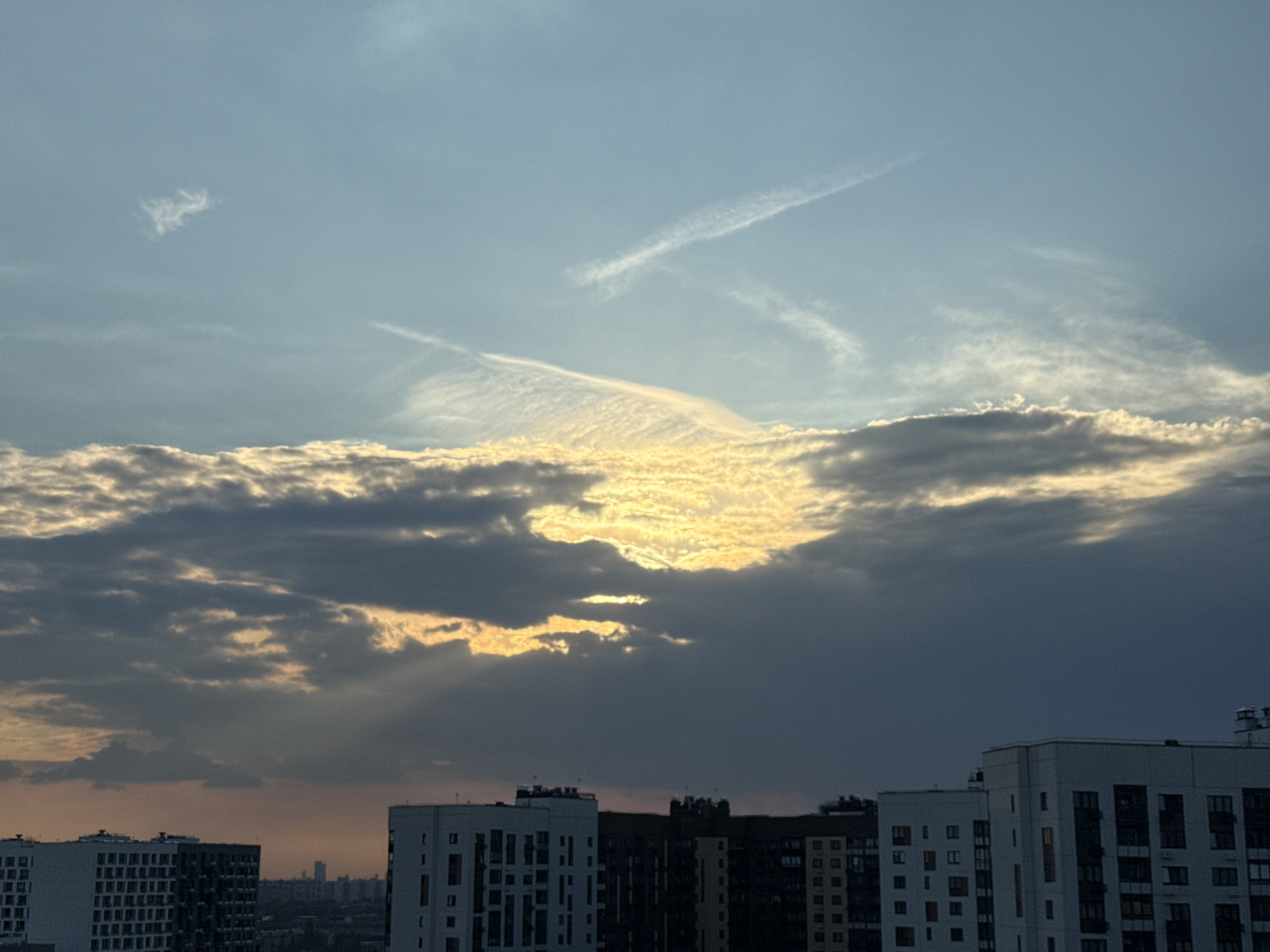 The image shows a cityscape in Lyubertsy, Russia. In the foreground are multiple modern apartment buildings with visible windows and balconies. The buildings range in color from white to black. Above the buildings, the sky is dominated by a thick layer of dark, gray clouds with bright, golden sunlight peeking through gaps in the clouds. The sun's rays are visible, shining downwards towards the buildings. The upper part of the sky is a lighter blue, with faint contrails and wispy clouds.
