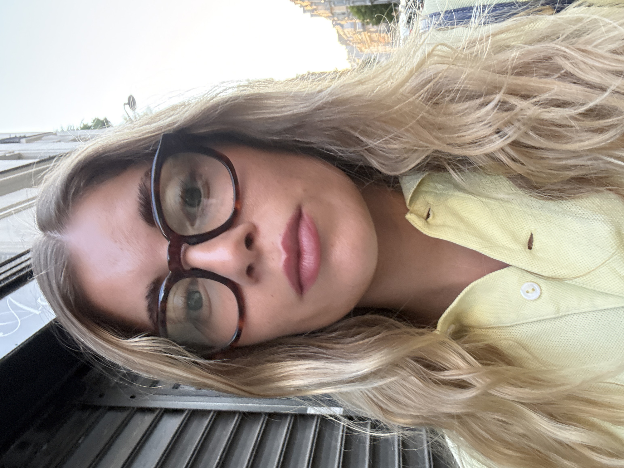 This is a close-up, head-and-shoulders selfie of a young Caucasian woman with blonde, wavy hair and light skin. She is wearing large, dark-rimmed glasses and a light yellow polo shirt. The background suggests an urban setting, with a grey metal shutter on the left and a building with balconies visible on the right, hinting at a European city street. The lighting is bright, with the sun low in the sky, suggesting it might be early morning or late afternoon. The woman is looking directly at the camera.