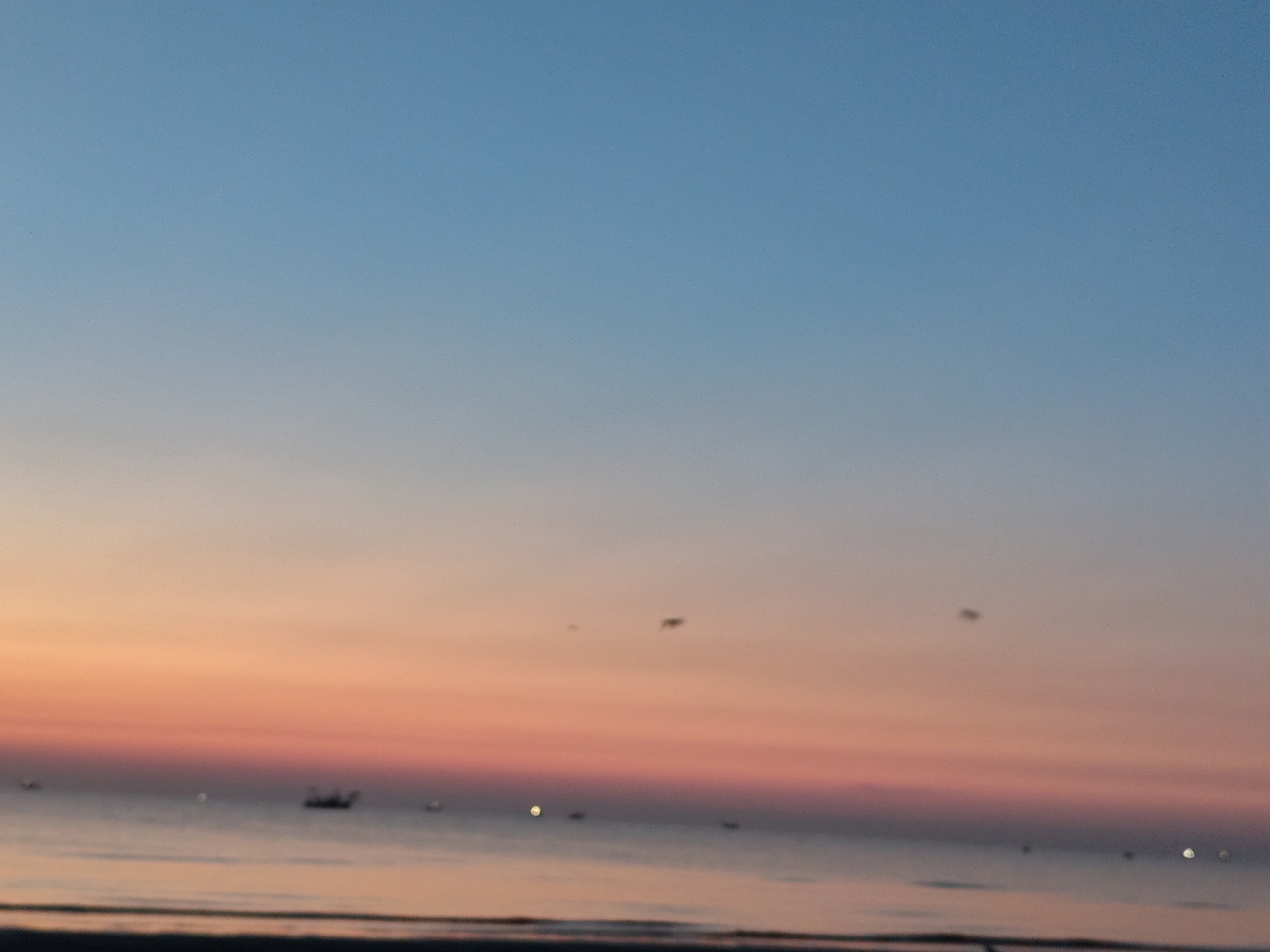 The image shows a hazy, blurred seascape with a gradient sky transitioning from light blue at the top to pink and orange near the horizon. There are a few indistinct dark shapes on the water, possibly boats, and some small, bright points of light that could be distant lights or reflections. The overall impression is of a sunset or sunrise over the sea. The location is identified as Rimini, Italy.