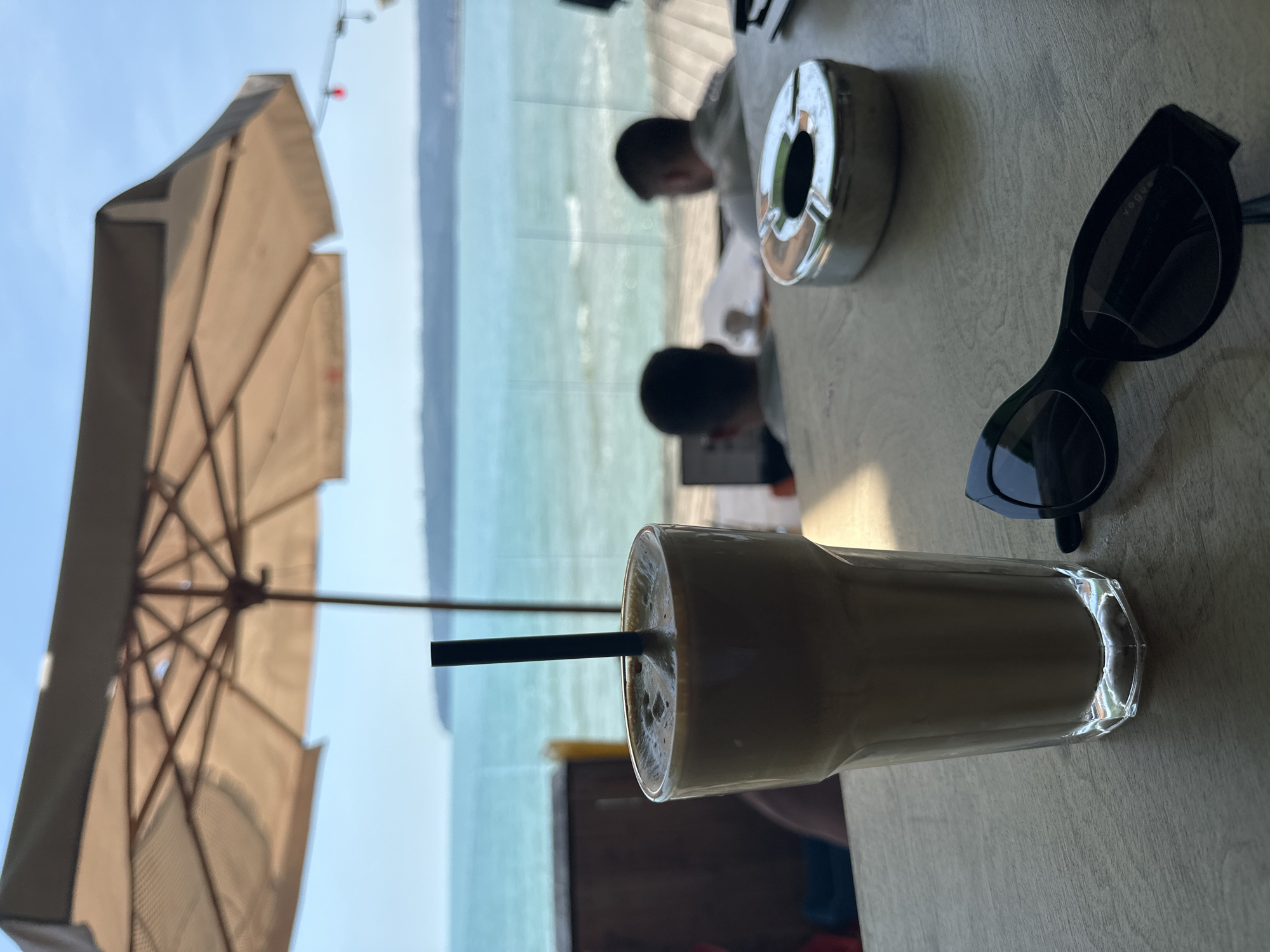 The image captures a first-person perspective from a wooden table in an outdoor or semi-outdoor cafe setting, overlooking a body of water. In the foreground, a tall, clear glass containing a light brown, frothy beverage with a black straw stands on the left. To its right, a pair of black, cat-eye style sunglasses with dark lenses rests on the table, with "Vogue" faintly visible on the right temple. Further right and slightly behind the sunglasses, a shiny, metallic, oval-shaped ashtray with three cigarette rests is positioned. The wooden table surface shows visible grain.
Beyond the table, through a glass barrier, two individuals are seated with their backs to the viewer, facing the water. The person on the left wears a dark top and is looking down, while the person on the right wears a lighter top. The background features a large, light-colored beach umbrella providing shade overhead, its internal structure visible. A string of faint red lights is discernible on the right side of the frame, near the horizon. The distant background reveals a vast expanse of turquoise-blue sea with small waves, meeting a light blue sky. A dark, blurred landmass or coastline is visible on the far horizon. The scene is well-lit, indicating daytime, with the foreground objects in sharp focus and the background increasingly blurred.