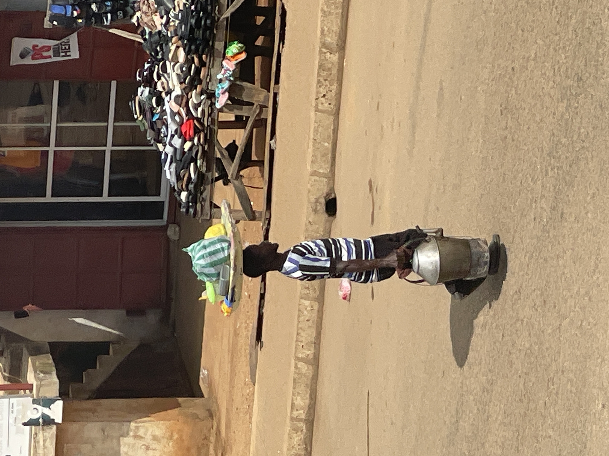 A young Black male walks across a sandy street, carrying a metal milk can in his right hand and a tray of goods balanced on his head. The boy wears a horizontally striped t-shirt and dark pants. The tray on his head contains a green and white striped bag, a yellow container, and other items. A table piled with shoes is visible in the background, along with a building with a red door and a window. The scene appears to be an outdoor market or street vendor area.