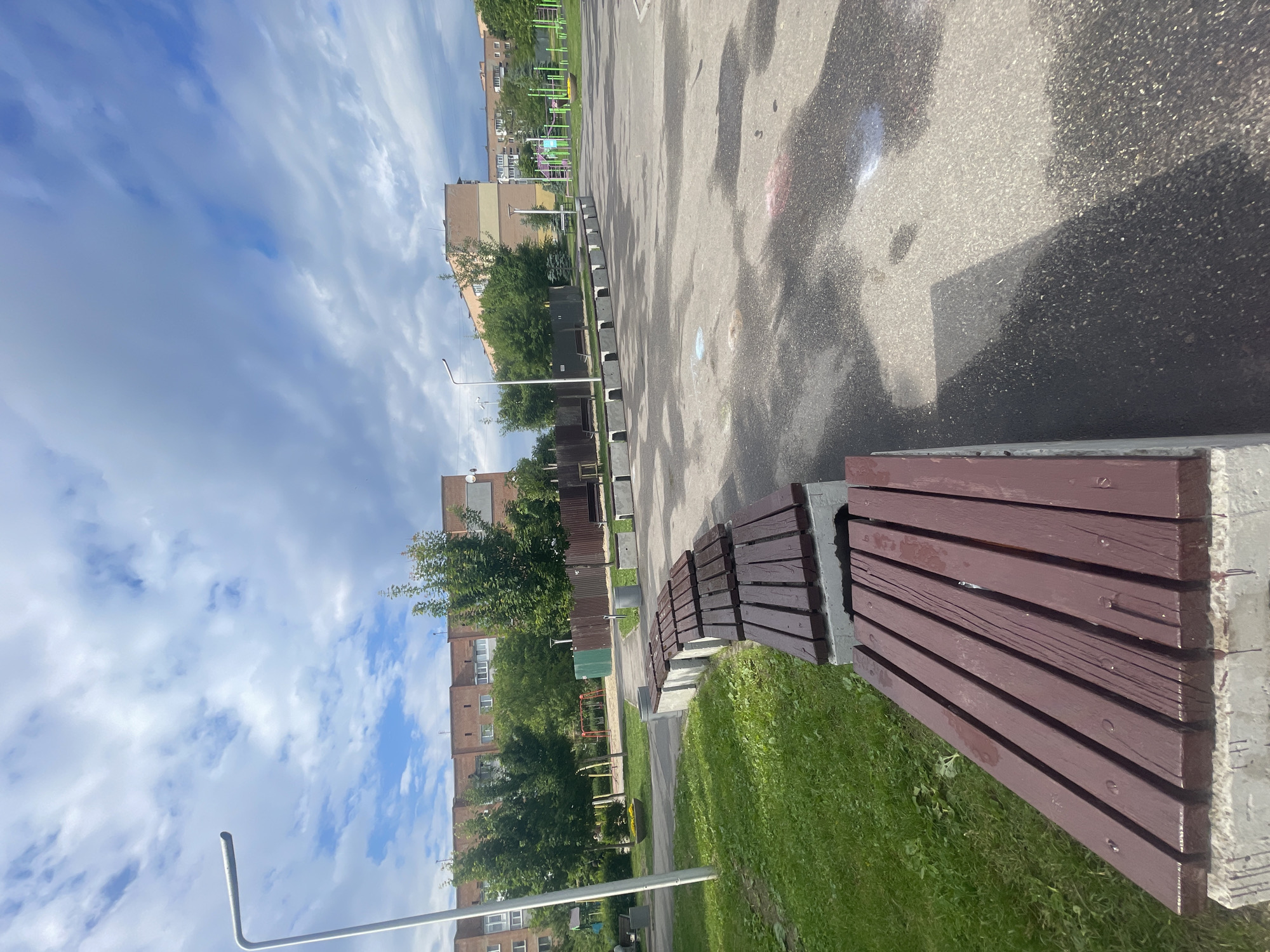 This image captures an outdoor urban or suburban scene in Poltava, Ukraine, on a bright, partially cloudy day. The perspective is from a low angle, looking forward along a paved area.
In the foreground and to the right, a series of dark brown wooden benches with light grey concrete bases are visible, situated on a strip of green grass. The paved asphalt area to the left of the benches appears damp with numerous wet patches, suggesting recent rain or watering. Faint, colorful marks, possibly chalk or paint, are visible on the asphalt near the foreground.
Further into the mid-ground, another line of concrete block structures, resembling bench supports without wooden slats, separates the paved area from an expanse of green grass and mature trees. Behind these trees, multi-story residential buildings with brick facades are visible, characteristic of post-Soviet architecture. Faintly discernible among the trees are elements of playground equipment, including slides and climbing structures, indicating a recreational space. Two metallic poles are present: a bent, silver pole in the bottom left foreground, and a taller streetlight further back on the left side of the paved area.
The sky, filling the upper portion of the image, displays a mix of blue patches and streaky white and light grey clouds. The overall impression is a quiet public space, likely a park or residential courtyard, with no people or specific activities taking place. No visible text is present in the image.