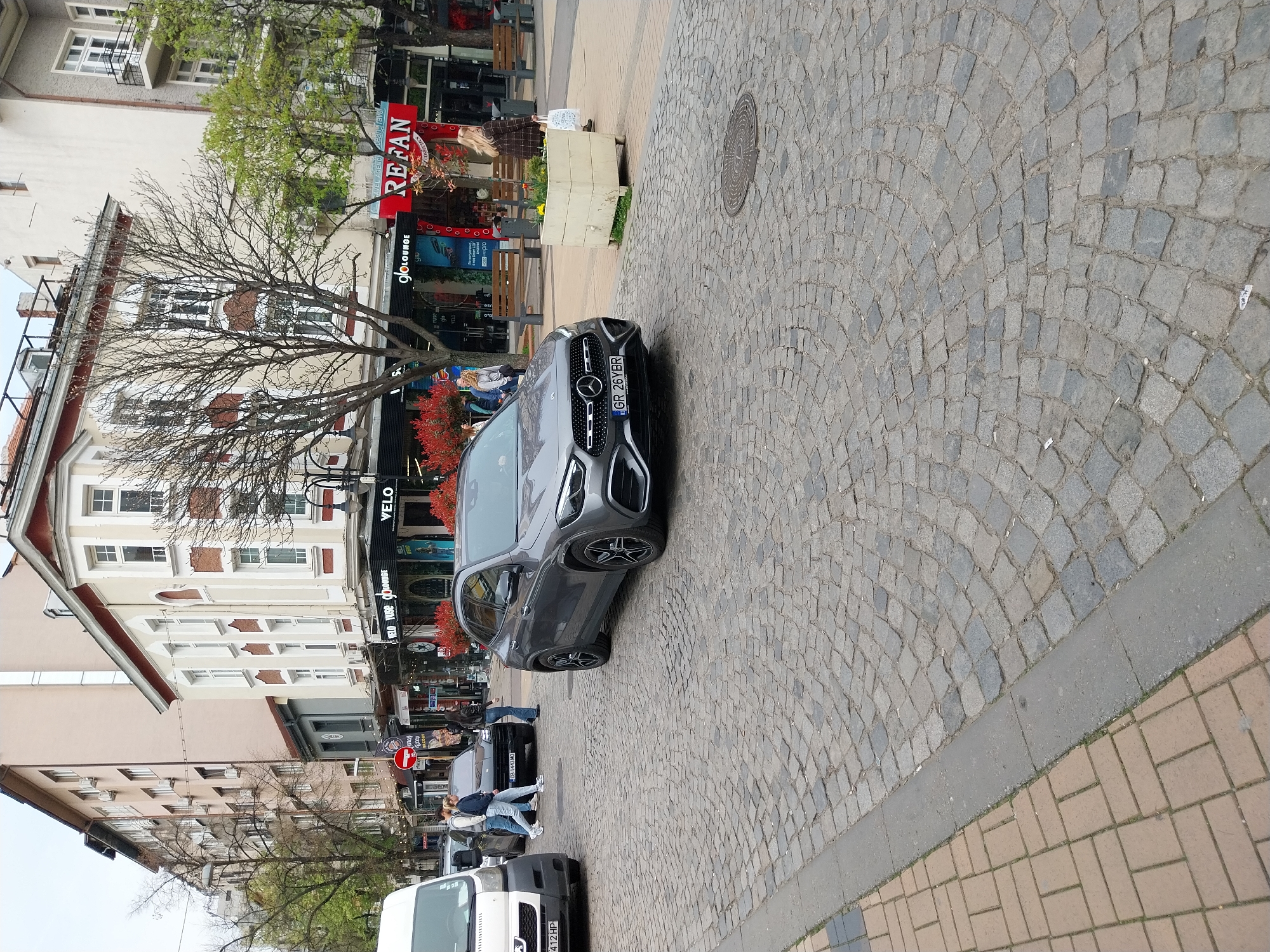The image captures a street scene in Sofia, Bulgaria. The focus is on a gray Mercedes-Benz parked on a cobblestone street. The car has a license plate reading "GR 26YBR." Further down the street, a dark-colored car with the license plate "CB 1443 MO" is visible, followed by a white van with the partially visible license plate ending in "412 HP." Pedestrians are walking on the sidewalk.
Buildings line the street. A "REFAN" store is prominently displayed with red signage. Other shops, including "gloLOUNGE" and "VELO VUSE VELO," are also visible. A bare tree is situated near the Mercedes-Benz.