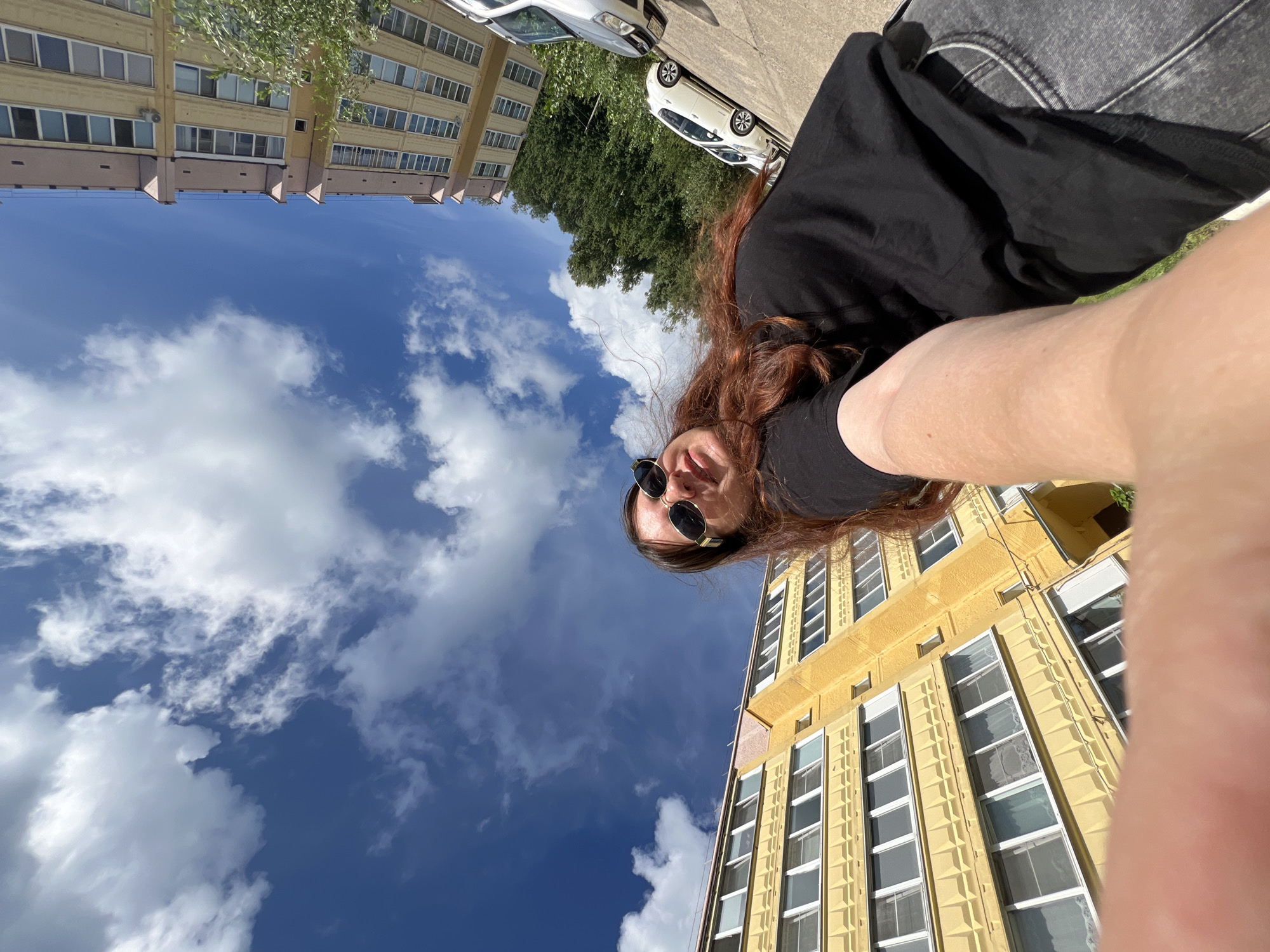 A selfie taken outdoors on a sunny day shows a woman with long, auburn hair wearing sunglasses and a black t-shirt. She is standing in front of a yellow apartment building with multiple floors and windows, and a light pink apartment building is visible in the background. The sky is blue with scattered white clouds. Trees and parked cars are also present in the scene. The image appears to be taken in an urban or suburban setting. Based on the context provided, the location is Noviy, Russia.