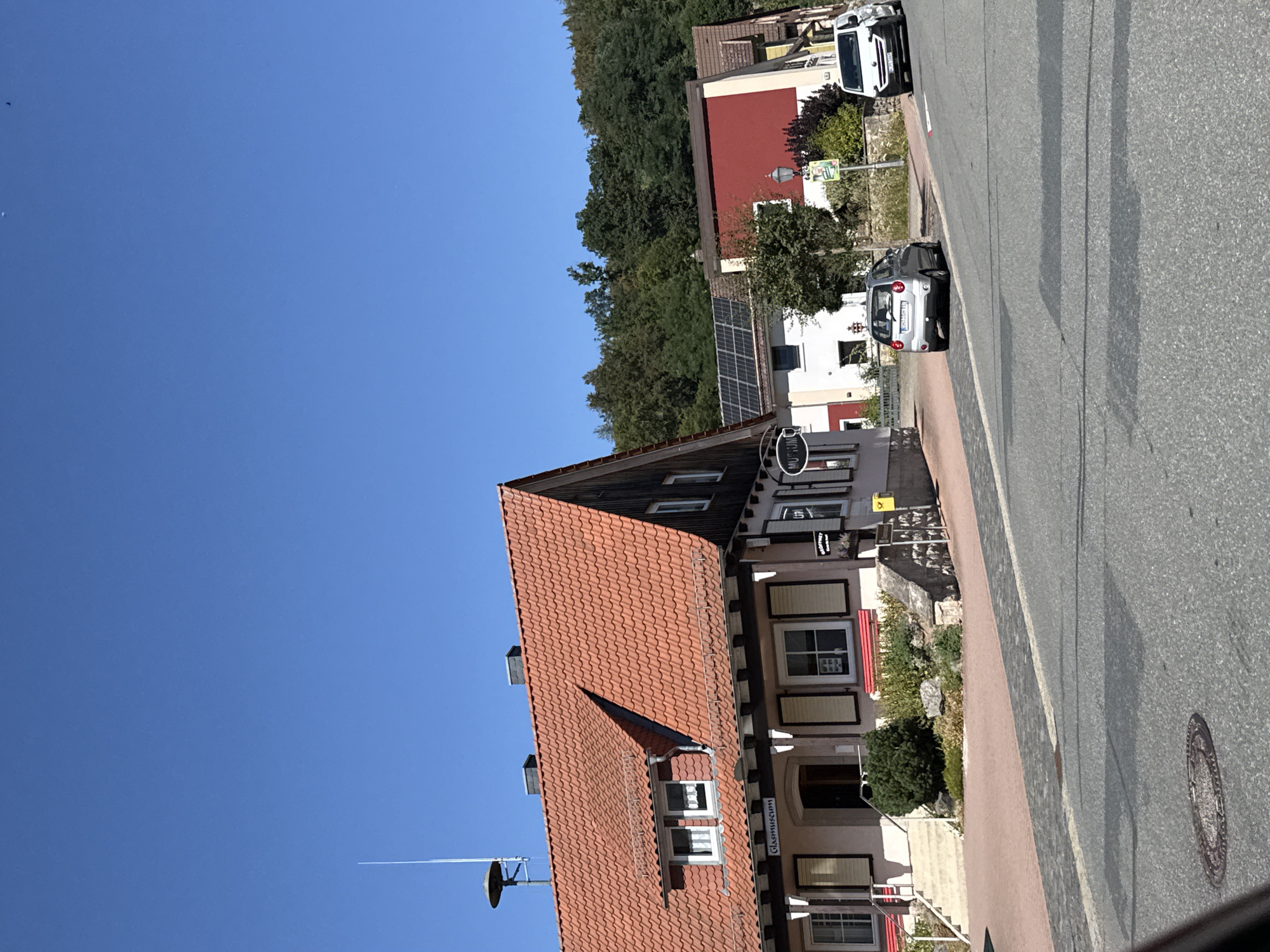 This is a daytime, eye-level, full-frame shot taken from a street in the town of Bad Sachsa, Germany, looking towards a street with buildings on the left and right. The sky is clear and blue. On the left side of the street is a two-story building with a red tile roof, a prominent mansard roof, and white trim. A sign above the entrance reads "Glasmuseum". There are several windows with shutters. In front of the building, a light-colored car is parked facing left. Further down the street, a white van is parked facing right. The street is paved with asphalt and has a cobblestone section on the left. Trees and vegetation line the background. The scene appears to be a quiet, residential or small town street on a sunny day.