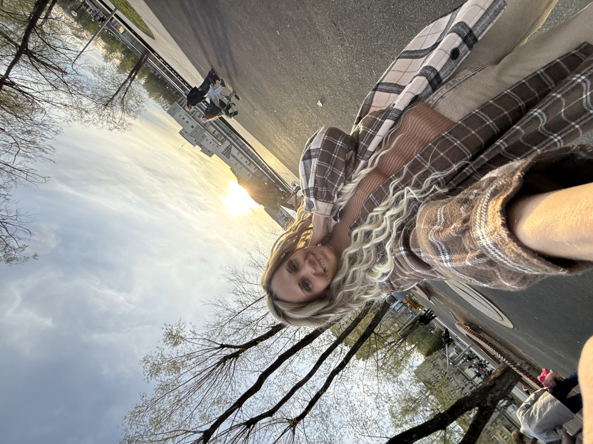 The image shows a woman with blonde curly hair, smiling at the camera. She is wearing a brown sweater and two plaid shirts, one brown and one tan. The photo appears to be a selfie taken outdoors, possibly in the city of Rusanovo, Russia, as suggested. The background includes trees with bare branches and a cloudy sky with a glimpse of the setting sun. In the distance, there are buildings, pedestrians, and a street. The photo seems to have been taken at an angle.