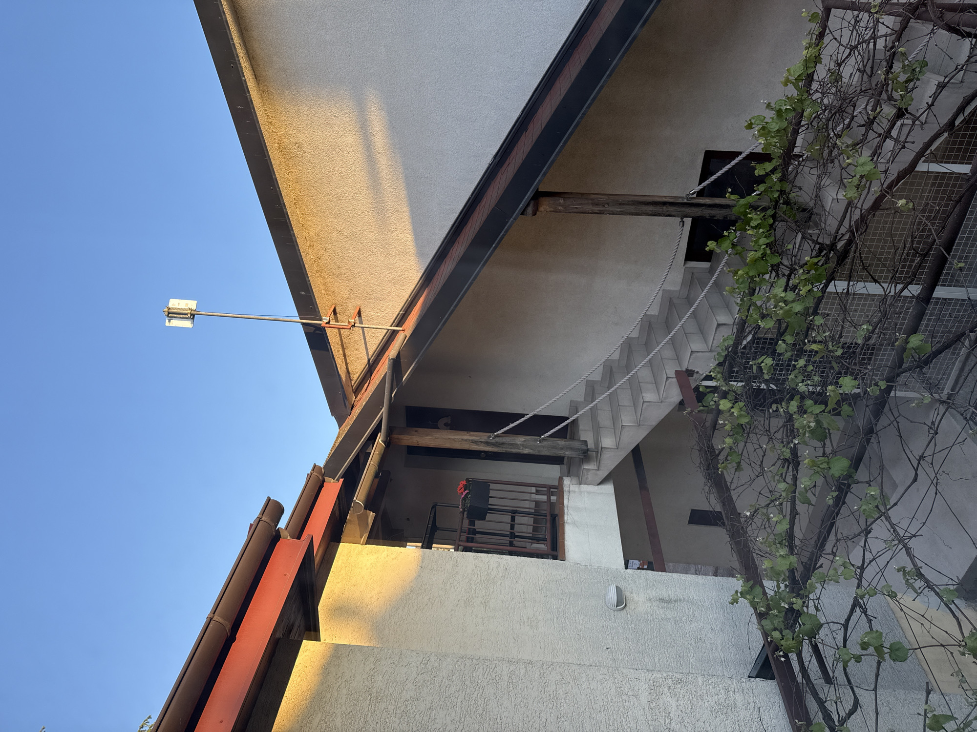 A low-angle shot looking upwards reveals a multi-level building in City of Zagreb, Croatia. The building is constructed of light-colored materials and features prominent horizontal lines. A large, flat roof extends outward, creating an overhang that casts a shadow. A staircase with white steps is visible beneath the overhang. To the side, a trellis with climbing vegetation adds a natural element to the scene. A street light extends from the building towards the clear, blue sky. A small balcony with a railing can be seen on one of the levels. The scene is bathed in warm light, suggesting it's either morning or late afternoon.