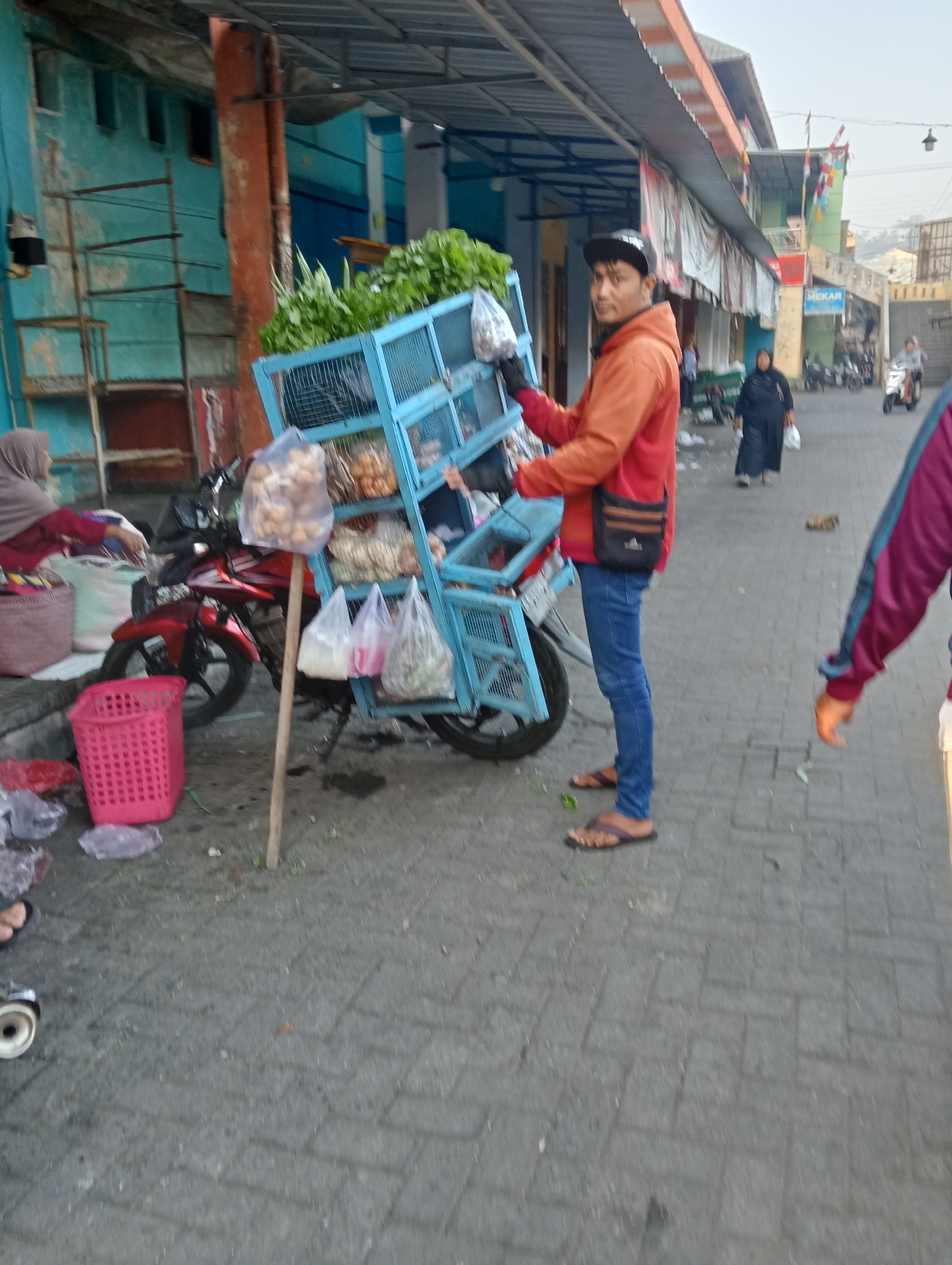 In Grabag, Indonesia, a street vendor stands next to a blue metal cart loaded with produce, including leafy green vegetables and bagged goods. The vendor, wearing an orange hooded jacket and blue jeans, has a black bag slung across his body. The cart is attached to a red motorcycle parked on a paved street. In the background, other people are visible, along with buildings and parked motorcycles. The scene appears to be a local market or street-side vendor area.