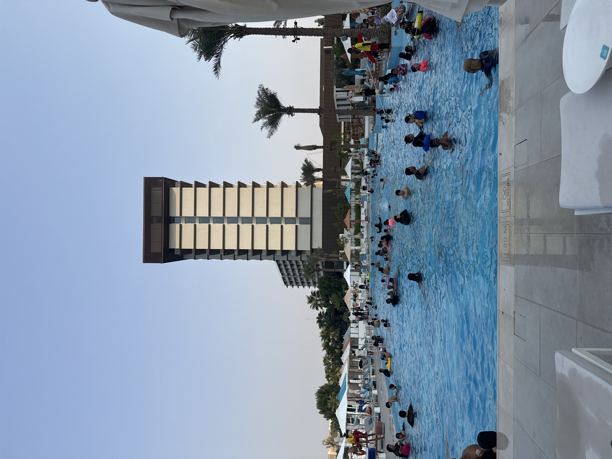 The image depicts a large, crowded outdoor swimming pool at a resort in the United Arab Emirates. The pool is filled with numerous people of various ages, many of whom are swimming, floating on inflatable rings, or standing in the water. The water is a vibrant blue, and the surrounding area features lounge chairs, umbrellas, and palm trees. In the background, a tall, modern building with a distinctive facade rises against a clear, pale blue sky. A lifeguard station with a red lifeguard on duty is visible on the left side of the pool. The overall scene suggests a lively and recreational atmosphere at a popular tourist destination.