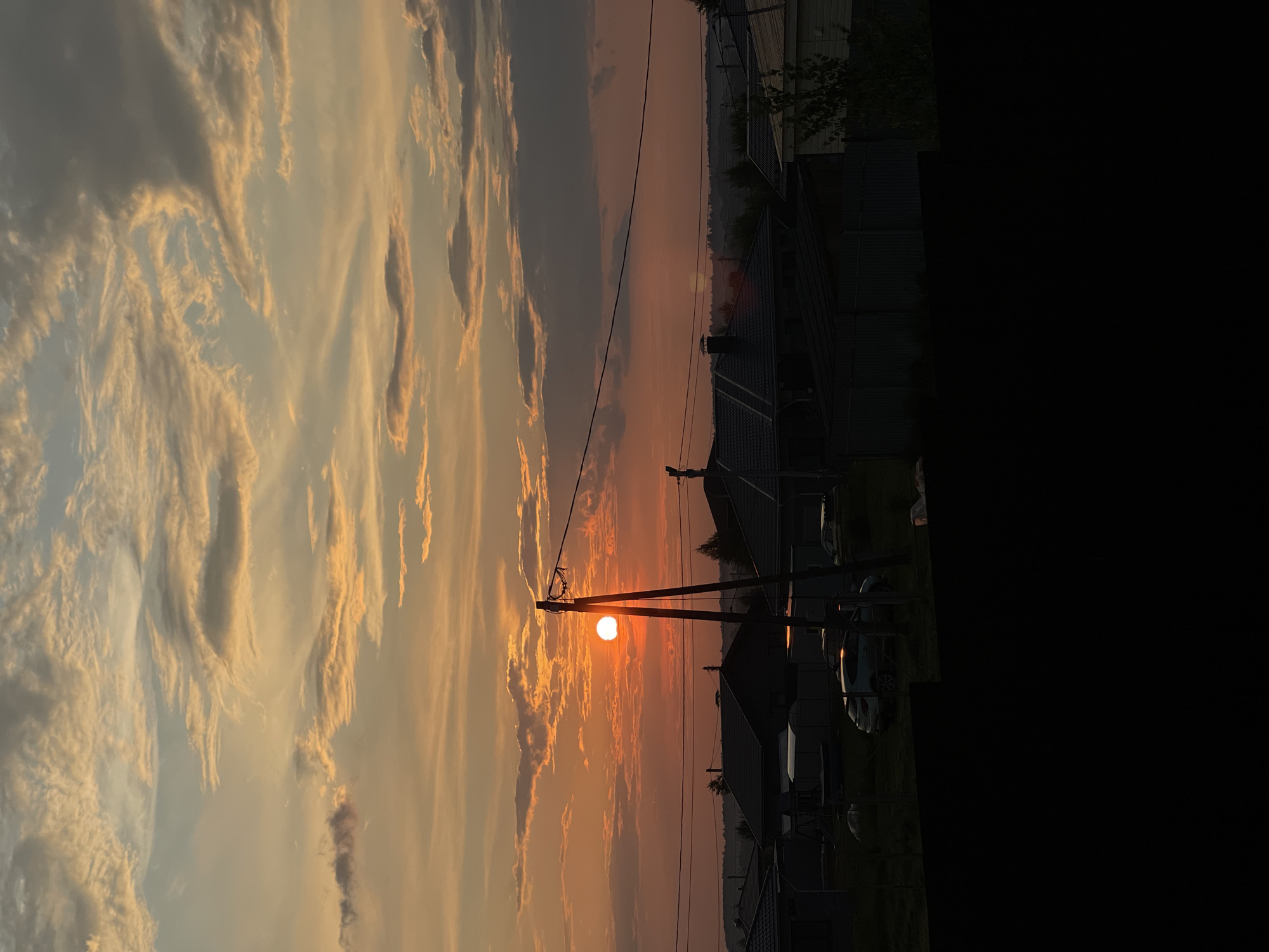 The image captures a sunset scene over a suburban area. The sky is filled with dramatic clouds, painted in hues of orange, yellow, and gray, with the sun a bright, distinct orb low on the horizon. In the foreground, silhouetted against the sunset, are houses with dark roofs, power lines stretched across the frame, and utility poles. A few cars are visible parked in the driveways or yards of the houses. The lower portion of the image is dominated by a dark, indistinct foreground, possibly a fence or landscaping. The overall impression is one of a tranquil, dusky evening in a residential neighborhood.