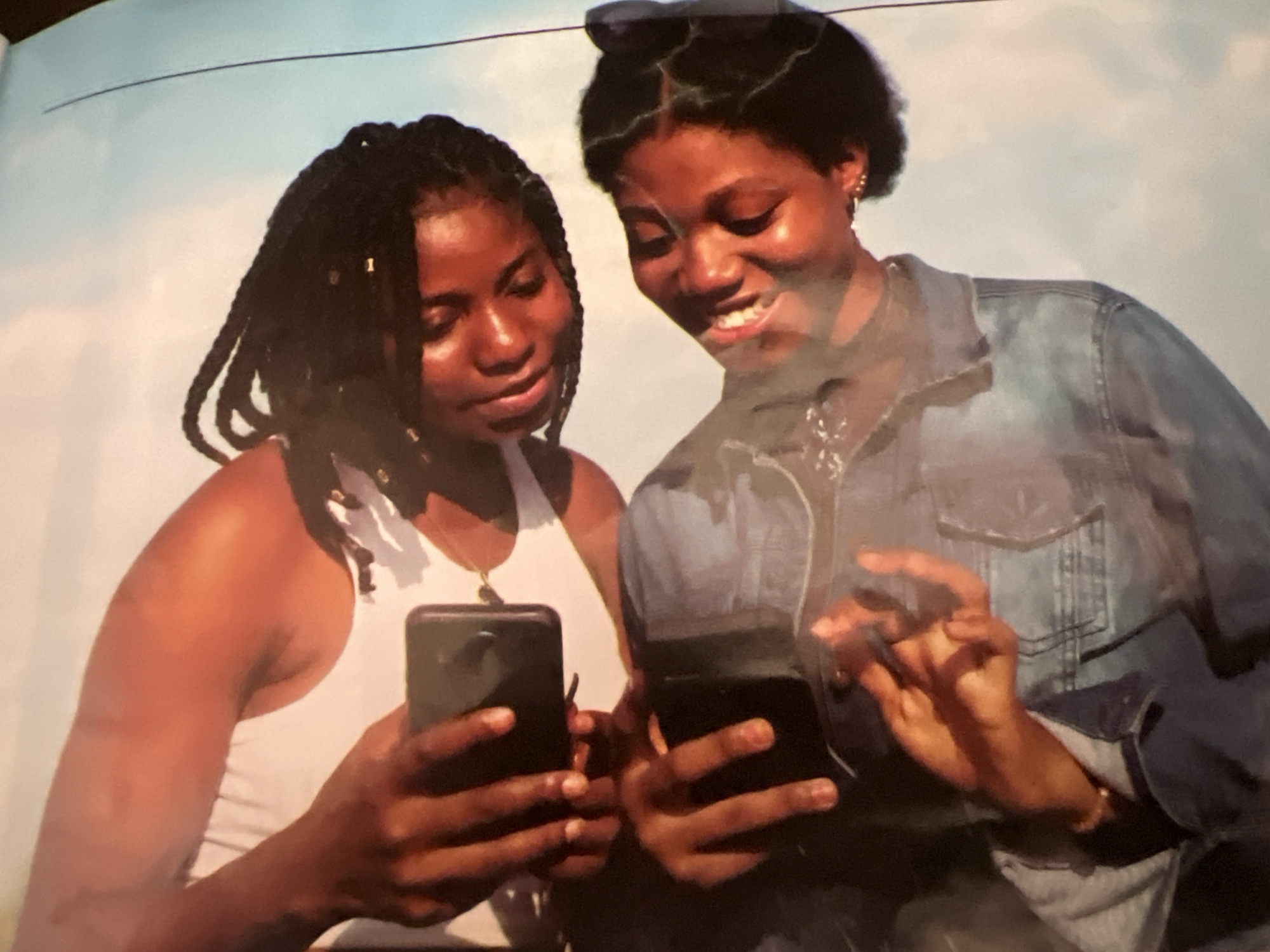This is a photograph of two young Black women, seemingly taken outdoors against a bright, possibly cloudy sky. The image is oriented vertically.
The woman in the foreground, appearing to be younger, is wearing a denim jacket and is looking down with her eyes closed and a wide smile. She has dark hair pulled up into a bun. Her right hand is visible, with fingers extended, and she is wearing a bracelet on her left wrist.
The woman behind her, who appears slightly older, is wearing a white tank top. She has dark hair styled in long braids. She is looking down and to her left, with her eyes closed and a gentle smile. She is holding a smartphone in her left hand, seemingly taking a selfie.
The overall impression is of a casual, happy moment shared between the two individuals. There is no identifiable location in the city of Jalingo, Nigeria, or any other specific urban setting evident from the background. The environment appears to be an open outdoor space.