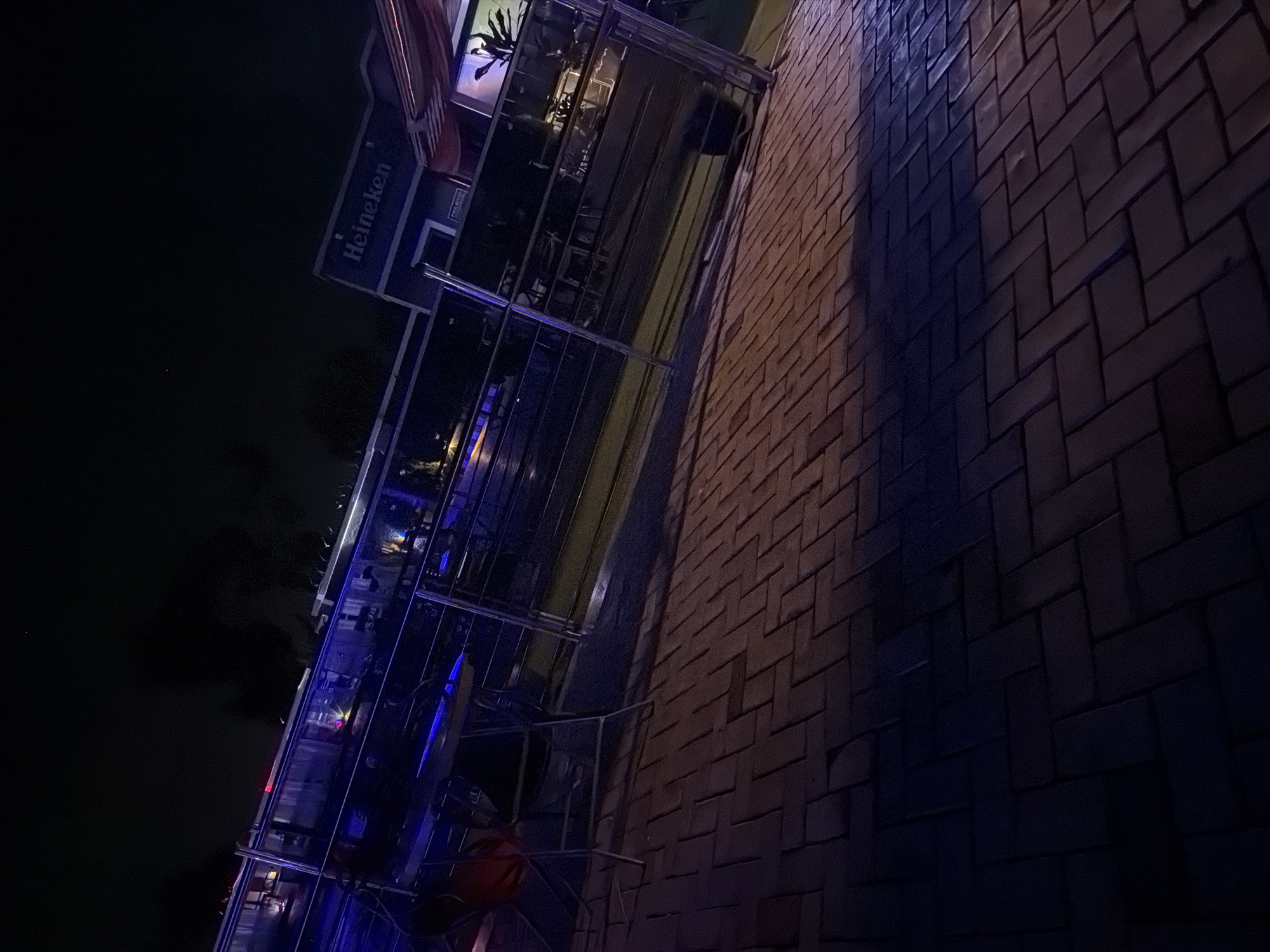 The image depicts an outdoor seating area at night, likely a restaurant or bar, with a prominent "Heineken" sign visible in the background. The ground is paved with brickwork, laid in a herringbone pattern. A metal railing separates the seating area from what appears to be a lower level or walkway. Blue and purple lighting illuminates parts of the scene, creating a dramatic ambiance. In the foreground, a table and chairs are visible, suggesting patrons were present or expected. The overall impression is one of a dimly lit, commercial establishment. There is no indication of the city of Bukuru, Nigeria within the image.