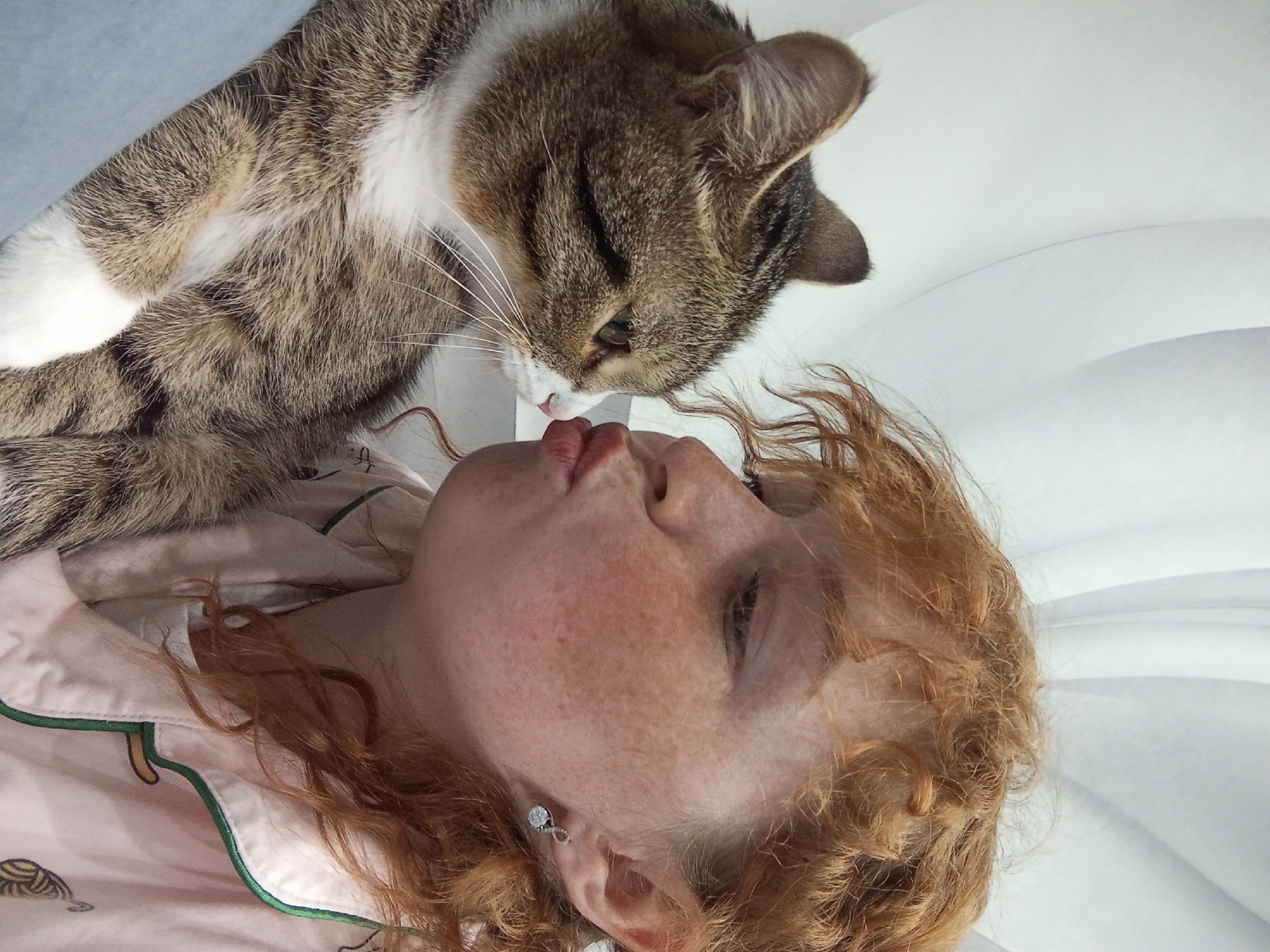 A woman with red, curly hair is lying on a white surface. Her head is tilted back, and her lips are slightly pursed. A tabby cat with white markings on its chest and paws is positioned above her face, with its nose close to her lips, possibly licking them. The woman is wearing light pink pajamas with a green trim and a pattern of turtles and mushrooms. The background is a white, slightly textured surface, possibly bedding. The photo appears to be taken indoors, likely a personal space. No location is evident.