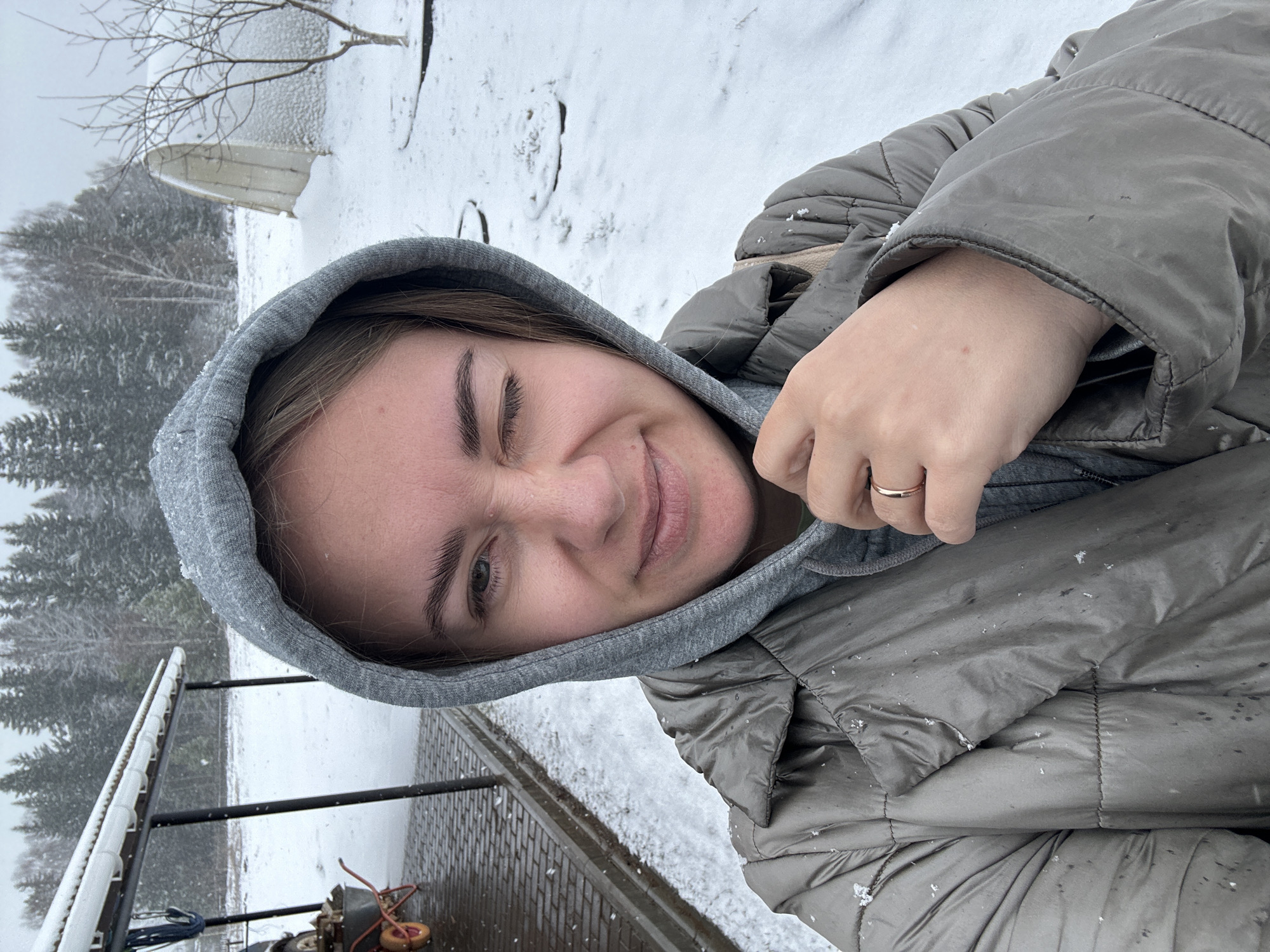 A young woman with brown hair and eyebrows is the primary subject of the image. She is wearing a gray hooded sweatshirt under a gray puffer jacket. Her right eye is closed in what appears to be a wink or squint. A simple gold ring is visible on her ring finger. The background suggests a snowy, outdoor setting. Trees and what appears to be a snow-covered boat can be seen in the distance. In the foreground, there is what seems to be a brick surface partially covered with snow. Scattered snowflakes are visible on the woman's jacket.
The image location might be Уральское, Russia.