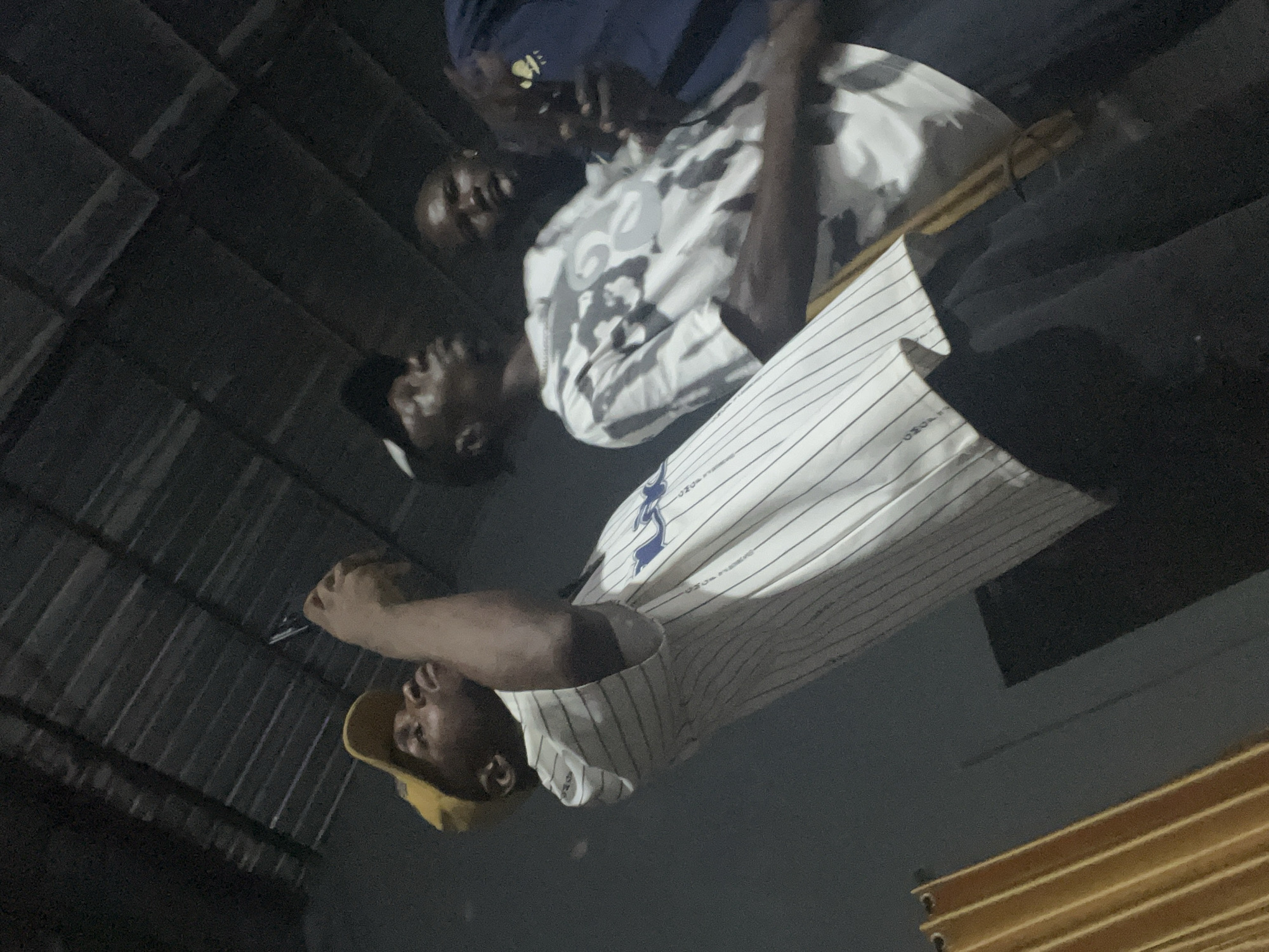 Three young Black men are gathered under a corrugated metal roof structure. The man on the left, wearing a yellow baseball cap and a white and blue pinstripe shirt, is holding up a smartphone, possibly taking a picture or video. The man in the middle wears a grey and white patterned t-shirt and looks upwards. The man on the right, wearing a dark blue shirt, is partially visible and appears to be interacting with the man in the middle. The background is dark and indistinct, with a yellow curtain visible on the left side of the frame. The scene suggests an informal gathering or event.