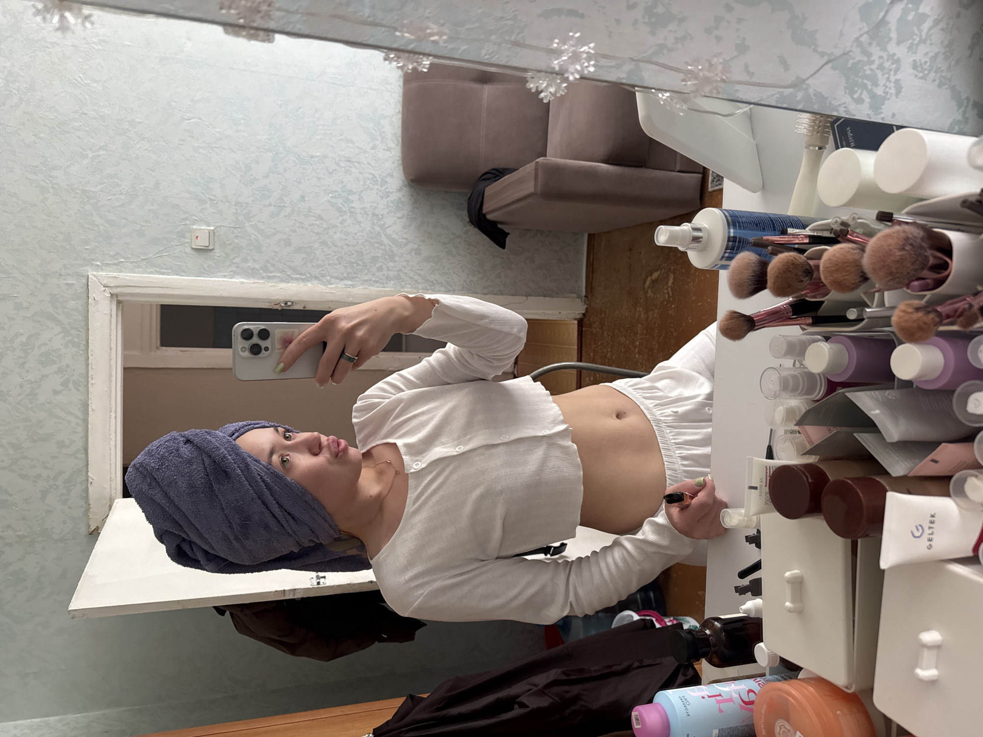 The image depicts a young woman taking a mirror selfie in an indoor setting, identified as the city of Chaikovsky, Russia. The woman is positioned centrally, facing her reflection in a large mirror that occupies the right side of the frame. She has a purple towel wrapped around her head and is wearing a white ribbed, long-sleeved, cropped top and matching white elastic-waist shorts. Her midriff is exposed. She holds a light-colored smartphone with three camera lenses in her right hand, visible in the upper center, directing it towards the mirror. Her left hand, holding a black pen or liner, rests on a white surface in front of her. She has light-colored nail polish on her fingernails and wears a dark ring on her left ring finger. Her lips are pursed, and her gaze is directed towards the phone.
In the immediate foreground, a white vanity table is cluttered with various cosmetic and skincare items. These include bottles of lotions and sprays (e.g., a blue bottle labeled "FOO," a clear bottle with "GELTEK"), an orange jar, several makeup brushes in containers, small white boxes with bow-shaped handles, and various tubes and vials.
The background shows a room with light blue patterned wallpaper. To the left of the woman, a white doorframe with a partially open white door reveals a dark interior and a person's dark-clad reflection. A dark jacket hangs on a hook to the far left. A light-colored power outlet or light switch is visible on the wall between the doorframe and the mirror. To the right of the doorframe, a light brown armchair with a dark cloth draped over it is visible. The floor appears to be light-colored wood or laminate. The mirror itself has decorative transparent snowflake-like elements on its right edge.