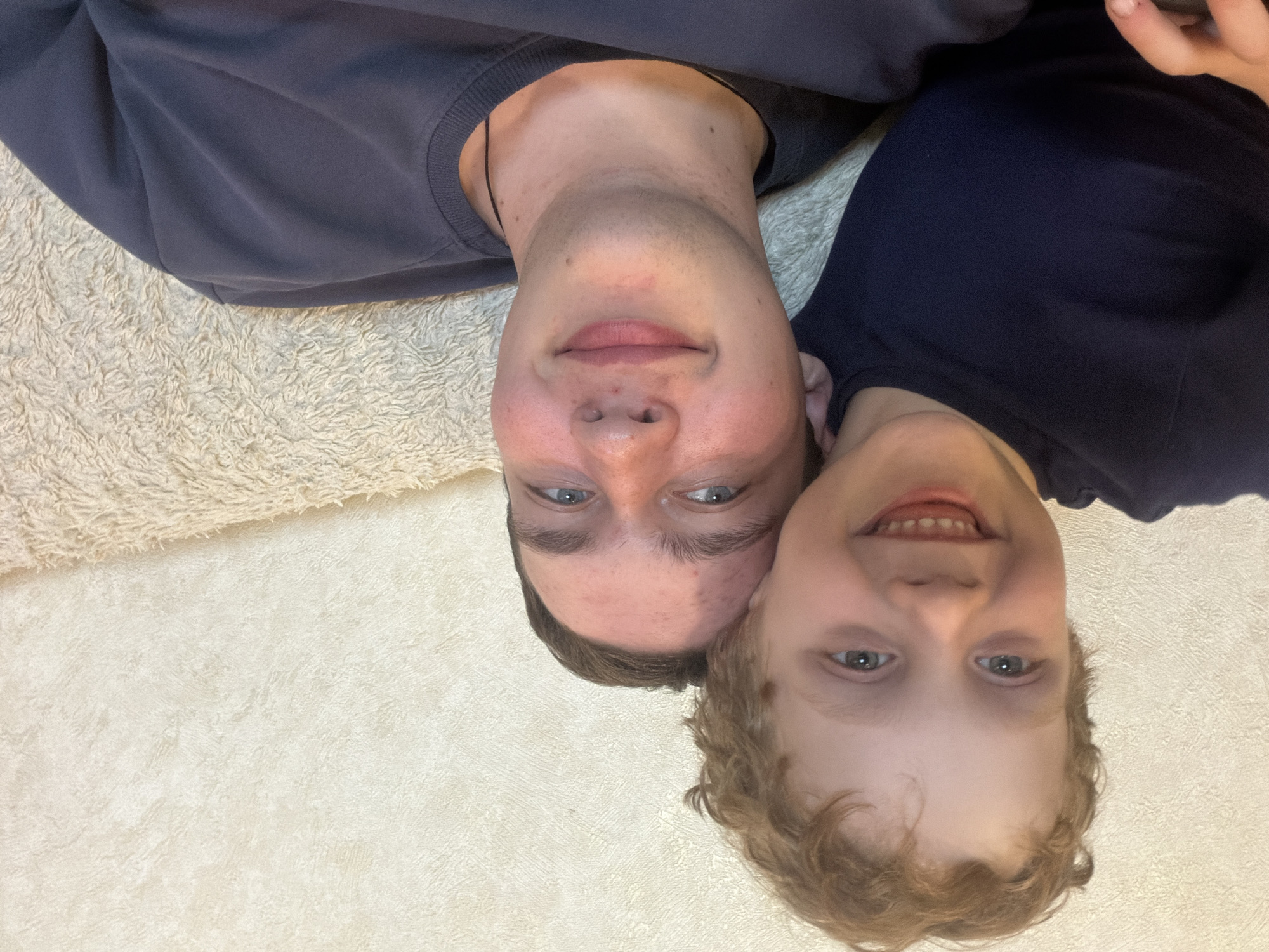 Two individuals, presumably a young boy and an older male, are positioned head-to-head and upside down in the frame. The younger individual has blonde, curly hair. Both are fair-skinned and have blue eyes. The older male has short brown hair and some acne. They are wearing dark-colored sweatshirts. The background is composed of a textured surface and a fluffy rug, suggesting an indoor environment, possibly a home. There is no verifiable information to conclude this image was taken in Severodvinsk, Russia.