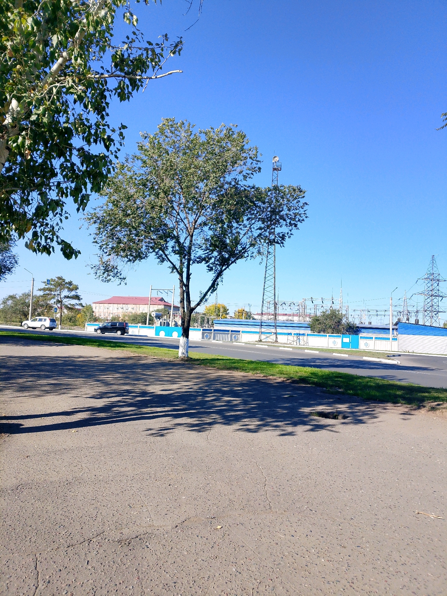 The image depicts an outdoor scene on a clear, sunny day, likely in the city of Kyzyl, Russia. In the foreground, a paved area, possibly a sidewalk or path, is visible with shadows cast by trees. Beyond this, a road runs horizontally across the frame. Two vehicles, a white SUV and a dark minivan, are driving on the road. On the other side of the road, there is a power substation with numerous electrical towers, wires, and buildings. A tall, slender radio tower is also present. Trees with green foliage line the area between the road and the substation. The sky is a vibrant, cloudless blue. The overall impression is one of an industrial area adjacent to a road, set against a bright, sunny backdrop.