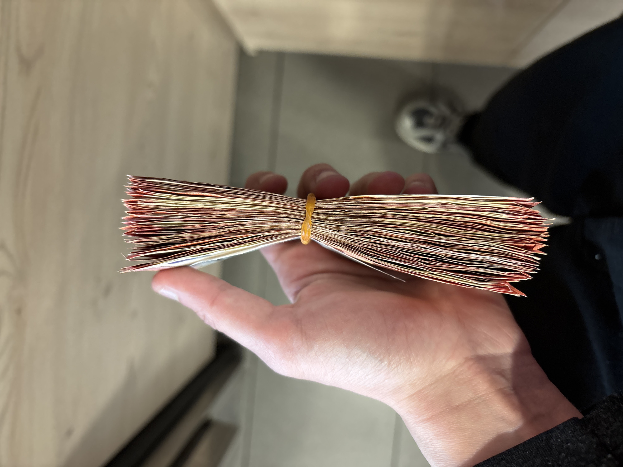 A hand holds a thick stack of what appears to be banknotes secured with a yellow rubber band. The denominations of the banknotes are not clearly visible, but there are hints of reddish-orange colors at the edges. The background features a light-colored wooden surface and a glimpse of flooring. A person is partially visible, wearing black clothing and a white shoe. The scene appears to be indoors. It is impossible to confirm the city of Sofia, Bulgaria based on visual information.