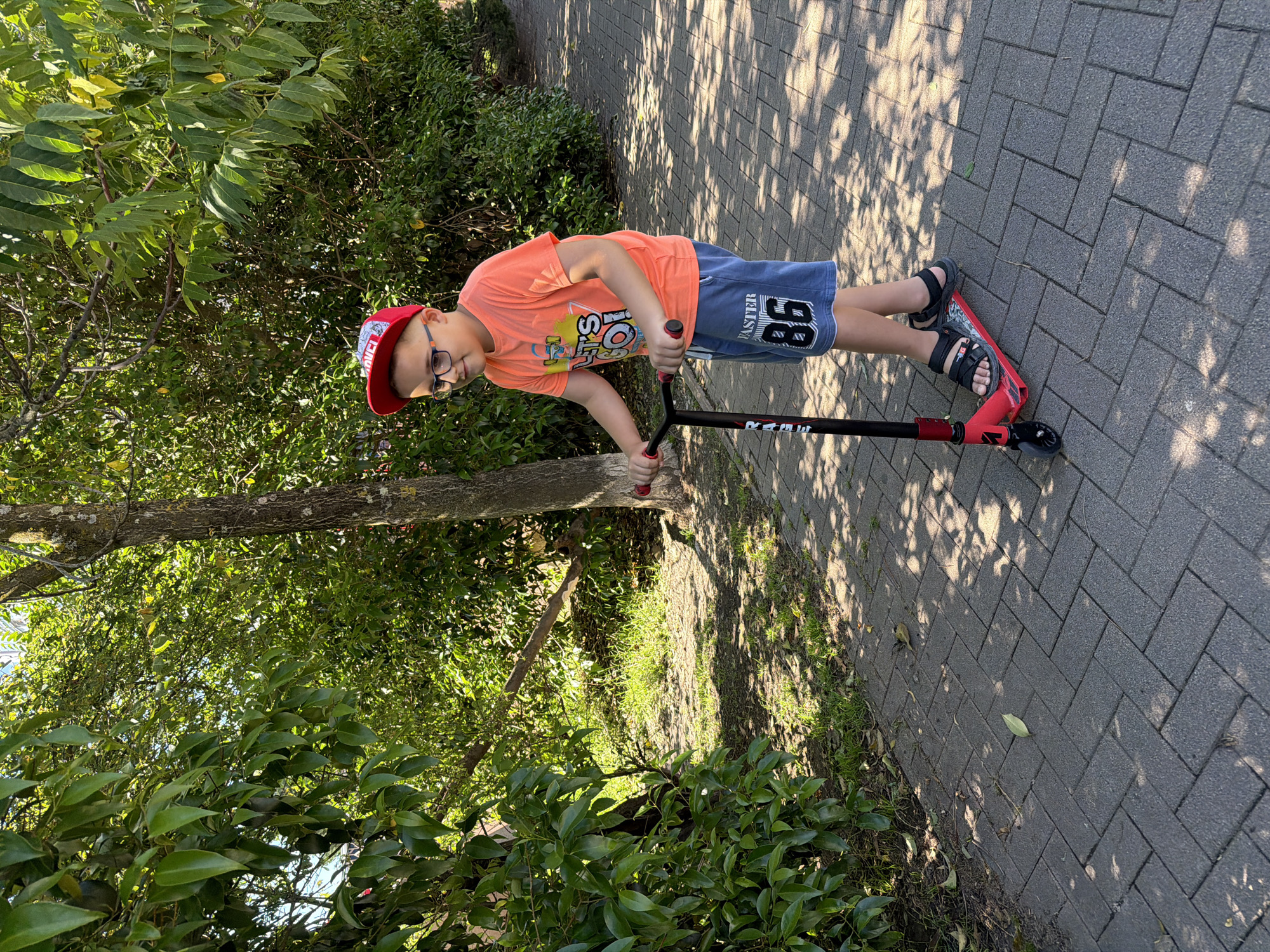 A young boy stands on a grey paved path, holding a red and black scooter. He is positioned towards the left side of the frame, looking directly at the viewer with a neutral expression. He wears an orange t-shirt, blue shorts, a red baseball cap, black sandals, and glasses.
Visible text on his clothing includes "MARVEL" partially seen on his cap, "LET'S PLAY!" on his t-shirt, and "FASTER 86" on his shorts. The scooter, which is red and black, has "RAGE" partially visible on its stem.
The setting is outdoors, likely a park or walking area, characterized by lush green foliage, including trees and bushes, bordering the paved path on both sides. Bright sunlight casts distinct shadows of tree leaves and branches onto the path, indicating it is daytime with clear weather. The ground is a dark grey interlocking paver path. The overall scene suggests a pleasant sunny day in an outdoor environment in Sochi, Russia.