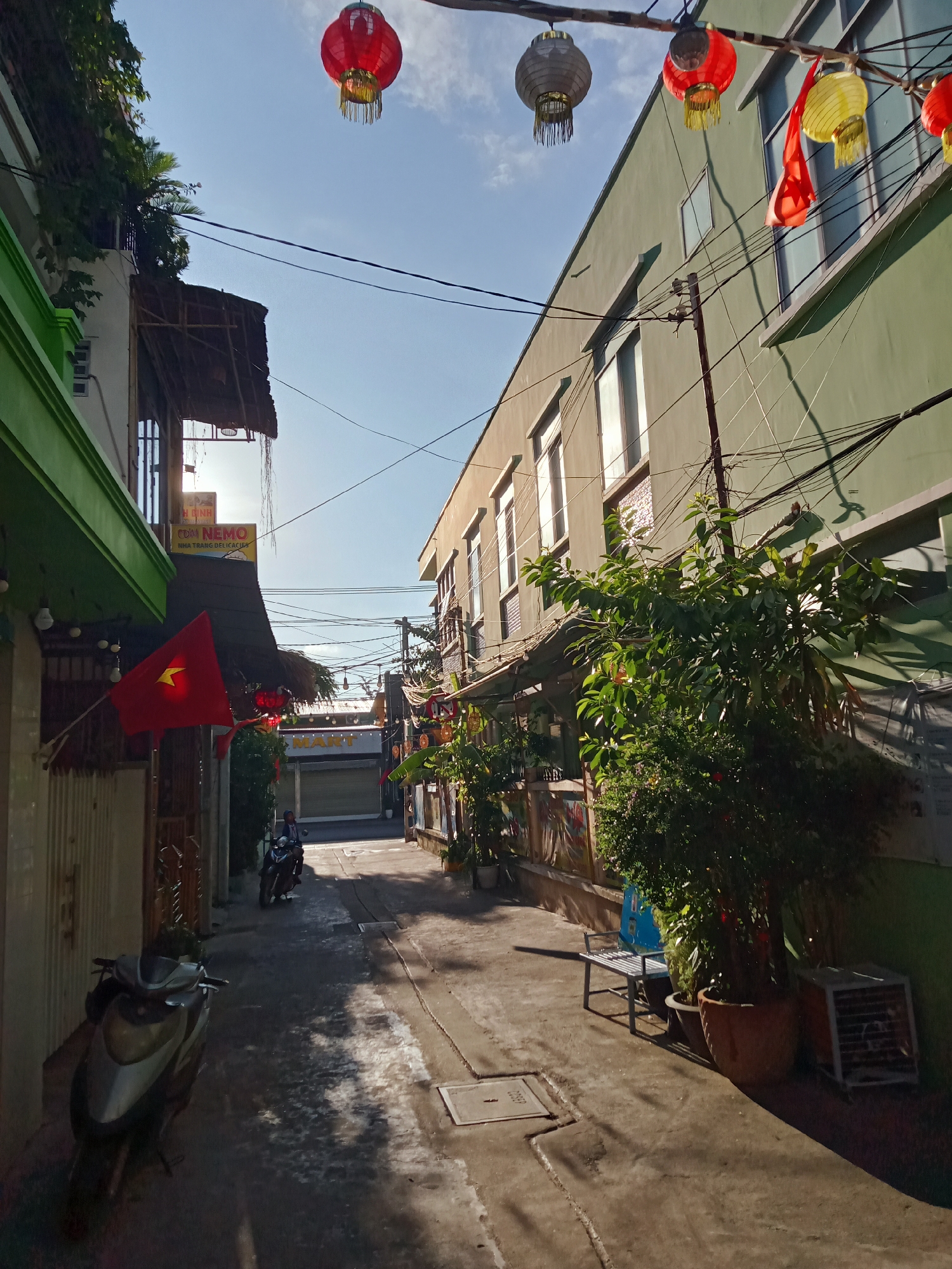 This image shows a narrow street in Nha Trang Ward, Vietnam. The street is lined with buildings on both sides. A Vietnamese flag hangs from a building on the left. Lanterns are strung across the street. A person sits on a parked motorcycle at the end of the street. Another motorcycle is parked on the left side of the street. There is dense overhead wiring in the street. Plants in pots, a metal bench, and an air conditioning unit are on the right side of the street.