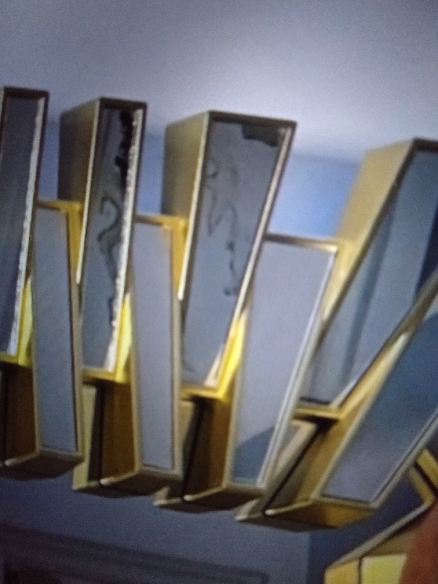 This is a close-up, somewhat blurry image of a decorative light fixture. The fixture appears to be made of gold-colored metal and clear or frosted glass panels. The panels are arranged in an angular, geometric pattern, creating a modern aesthetic. Some of the glass panels reflect the surrounding environment, showing hints of a blue wall and possibly other parts of the room or ceiling. There are also some dark smudges or reflections visible on the surfaces of some of the panels, possibly from dust or a manufacturing defect. The lighting in the image is not very bright, suggesting it might be evening or the fixture itself is not illuminated. The overall impression is of a stylish, contemporary decorative element. There is no visible text in the image. The setting is indoors, and the location context of Jalingo, Nigeria, doesn't provide specific visual cues from the image itself.