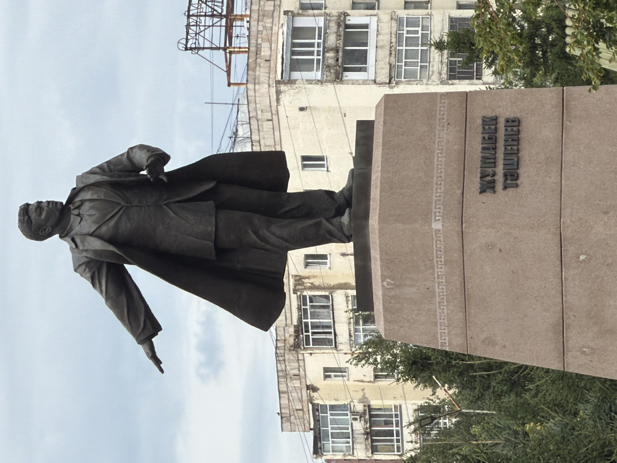 A full-length bronze statue of a man in a suit and overcoat stands on a stone pedestal. The man is facing left with his right arm extended, and his left arm is bent at the elbow. The pedestal has inscriptions in Cyrillic script that read "ЖУМАБЕК ТЭШЕНЕВ". In the background, there are multi-story apartment buildings and some industrial structures. Trees and greenery are visible in the foreground and middle ground. The sky is overcast. This monument is located in Astana, Kazakhstan.