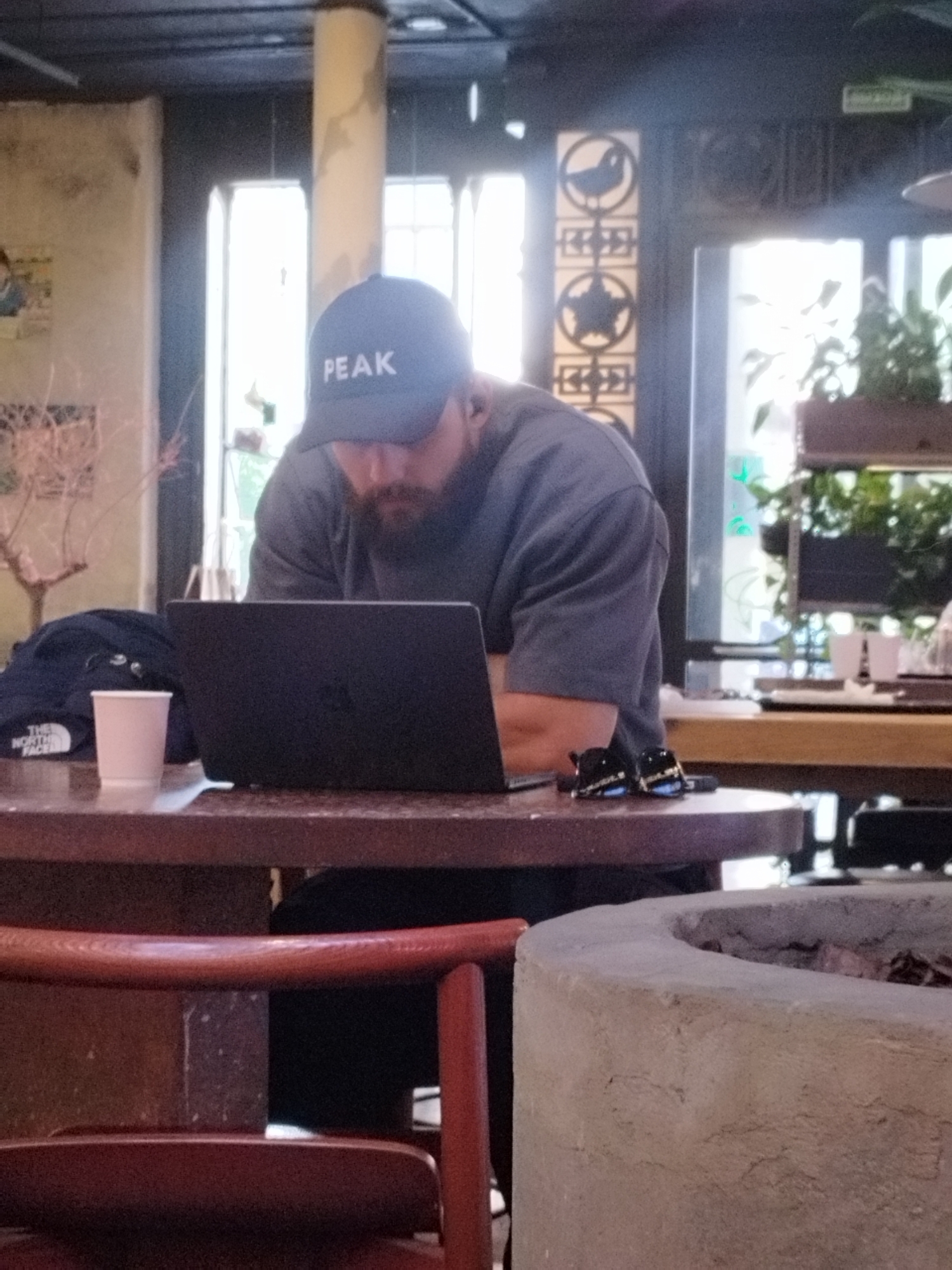 The image shows a man wearing a dark baseball cap with the word "PEAK" in white letters. He has a beard and is wearing a dark gray t-shirt. The man is seated at a dark brown round table, looking down at a black laptop. To the left of the laptop is a small pink cup and a dark blue jacket. On the table, to the right of the laptop, are a pair of black sunglasses. The setting appears to be indoors, possibly a cafe or restaurant, with large windows in the background. Through the windows, there is greenery and some decorative elements on the wall, including a circular motif with a bird and a star. The lighting suggests it could be daytime, perhaps late afternoon, given the indirect light coming through the windows. The overall mood seems calm and focused, with the man engrossed in his work or activity on the laptop. The presence of a chair in the foreground suggests a seating area within the establishment.