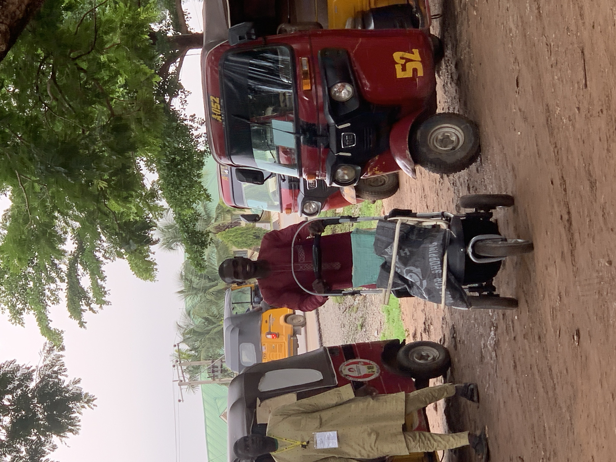 Two men are standing near three auto-rickshaws in what appears to be a parking lot or informal marketplace. The man on the left is wearing a light-colored traditional tunic and trousers with a yellow lanyard and badge around his neck. He is standing beside a red and black auto-rickshaw with a yellow side panel. The man on the right is wearing a dark red traditional tunic and light blue trousers. He is smiling and pushing a baby stroller, which has a black bag on it. Behind him are two more auto-rickshaws; one is red with the number "52" on its side, and the other is yellow. The ground is dirt, and there are trees and buildings in the background. Power lines and poles are visible above the trees. The lighting suggests an overcast day. The scene is likely outdoors in a busy area within Abuja, Nigeria, given the presence of auto-rickshaws, which are common modes of transportation there.