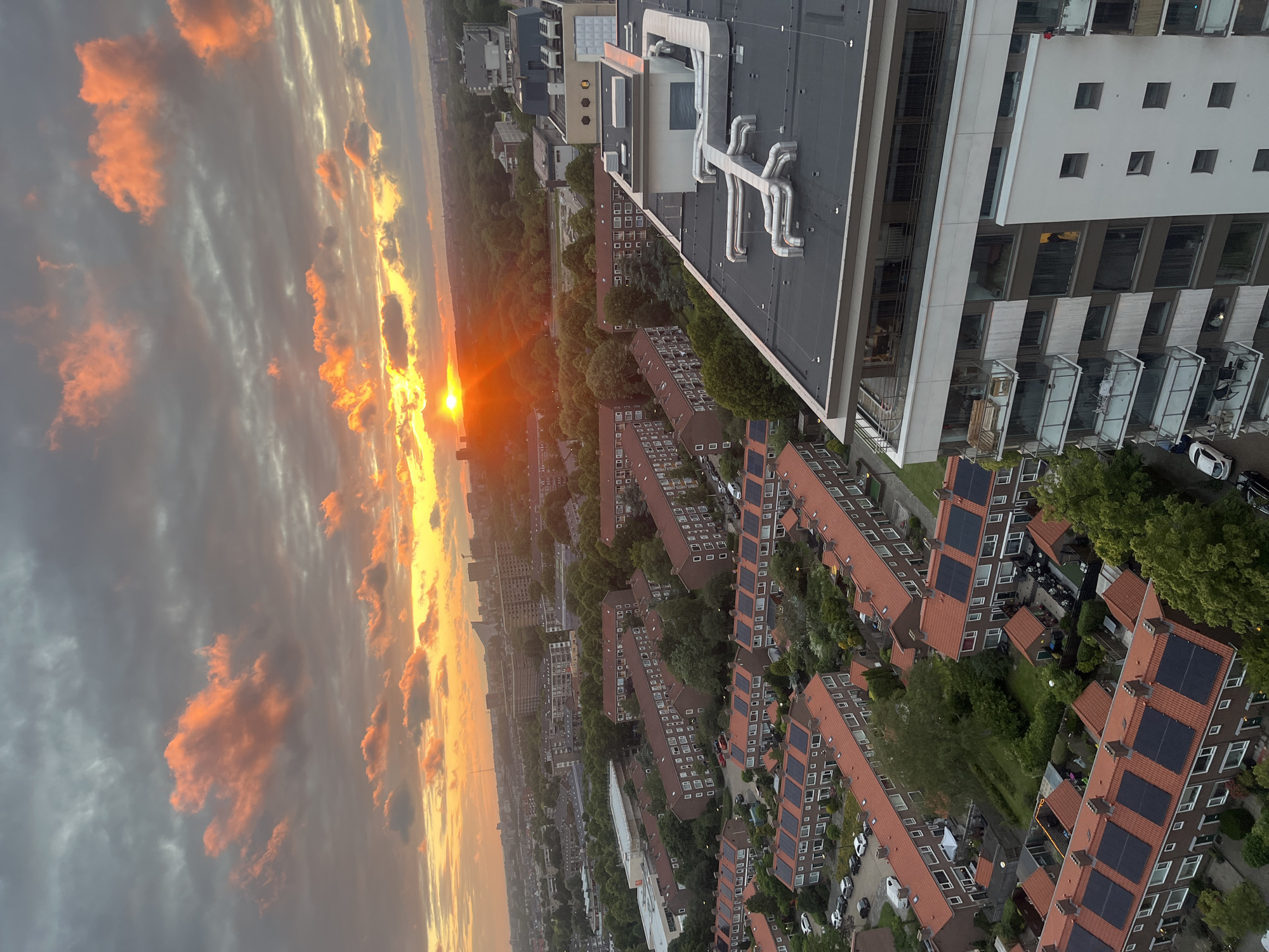 An aerial view captures a city skyline at sunset, with the sun low on the horizon, casting long shadows and illuminating the sky with vibrant orange and pink hues. A dense arrangement of residential buildings, characterized by red-tiled roofs and apartment complexes with balconies, stretches across the landscape. Some rooftops are equipped with solar panels. A modern, multi-story building with white and glass facades stands prominently in the foreground. The sky is filled with dramatic clouds, some tinted pink and orange by the setting sun, contrasting with darker, grayer clouds. Trees are scattered throughout the urban environment, adding patches of green amidst the architecture. The scene suggests a European city.