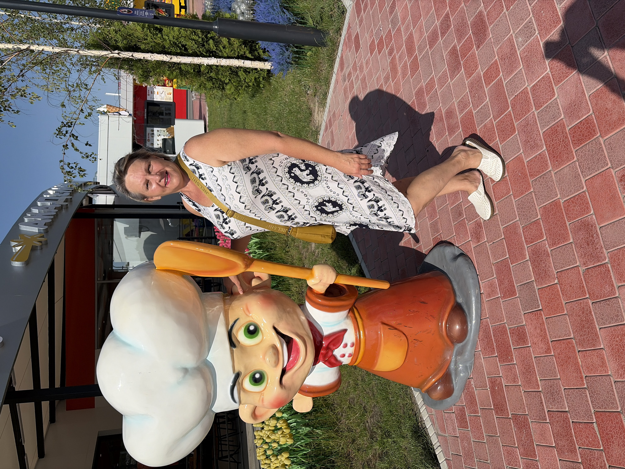 A middle-aged woman stands next to a cartoonish chef statue outside a building. She is wearing a sleeveless white dress with a black elephant pattern and a yellow crossbody bag. The statue is of a young boy wearing a chef's hat and apron, holding a large spatula. The background shows a building with a red and white facade, some trees, and a blue sky. The ground is paved with red brick tiles.