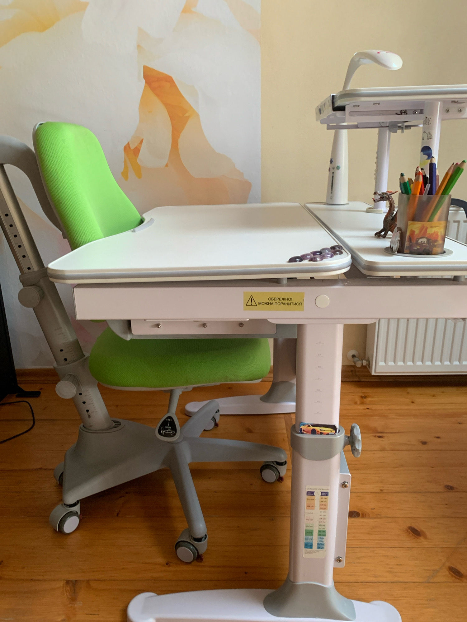 The image shows a child's ergonomic desk and chair set in a room with wooden flooring. The desk is white with pink accents and has an adjustable lamp and a pencil holder with colored pencils and a small dinosaur figurine. A yellow warning sticker with Ukrainian text is affixed to the desk's support. The chair is green and grey, with wheels. The background features a wall with a floral wallpaper pattern. There are no people present in the image. The image does not provide any information that would allow for the determination of its location in Kolomyia, Ukraine.