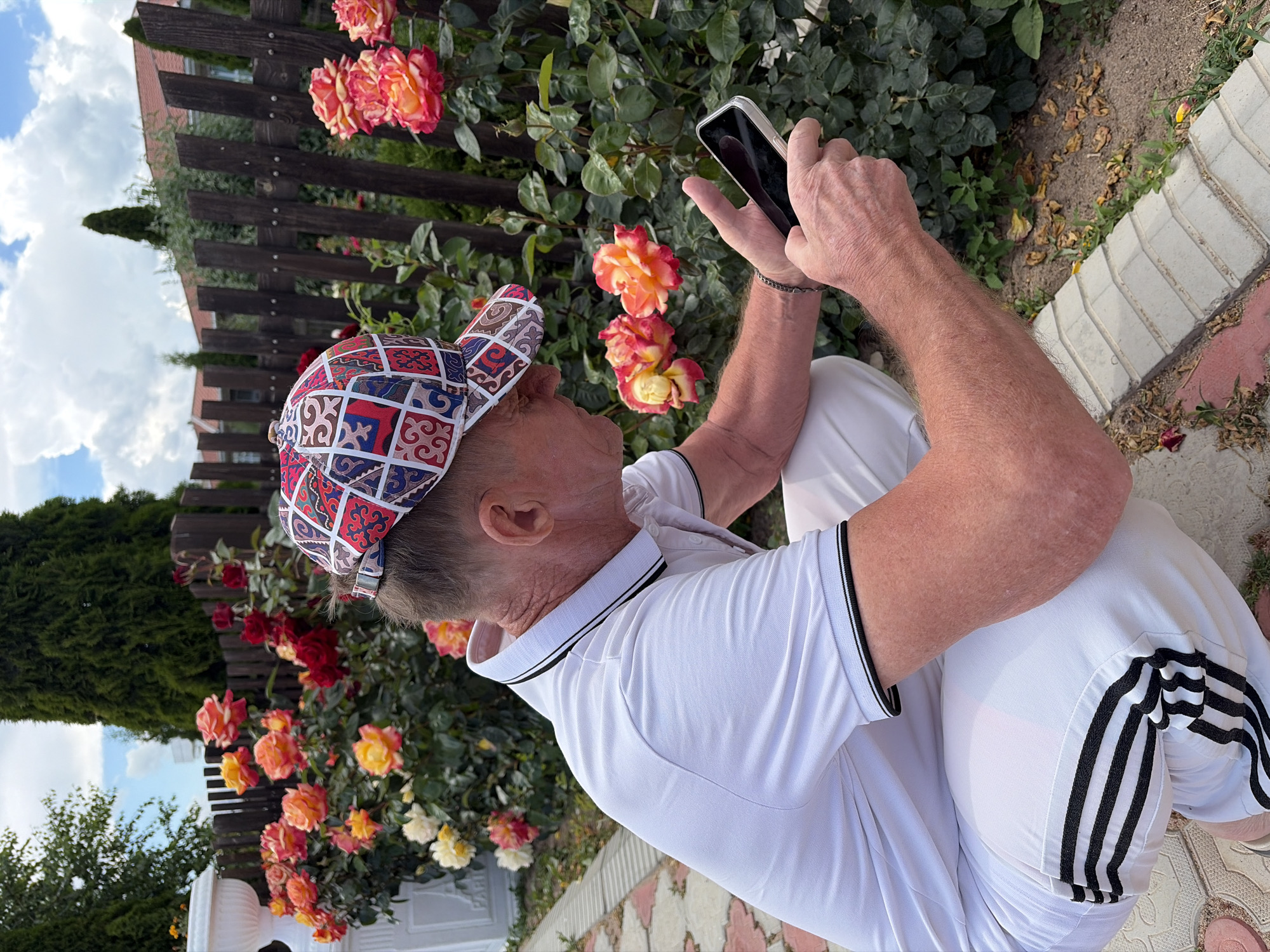 The image shows a man wearing a white polo shirt and a colorful patterned cap, taking a picture with his cell phone. He is outdoors, near a rose bush with vibrant red, orange, and yellow flowers. Behind him, a wooden fence separates the garden from a building. The sky is visible with some clouds, suggesting it is daytime. The man appears focused on capturing an image of the roses. The setting seems to be a residential garden or yard. There is no visible text in the image.